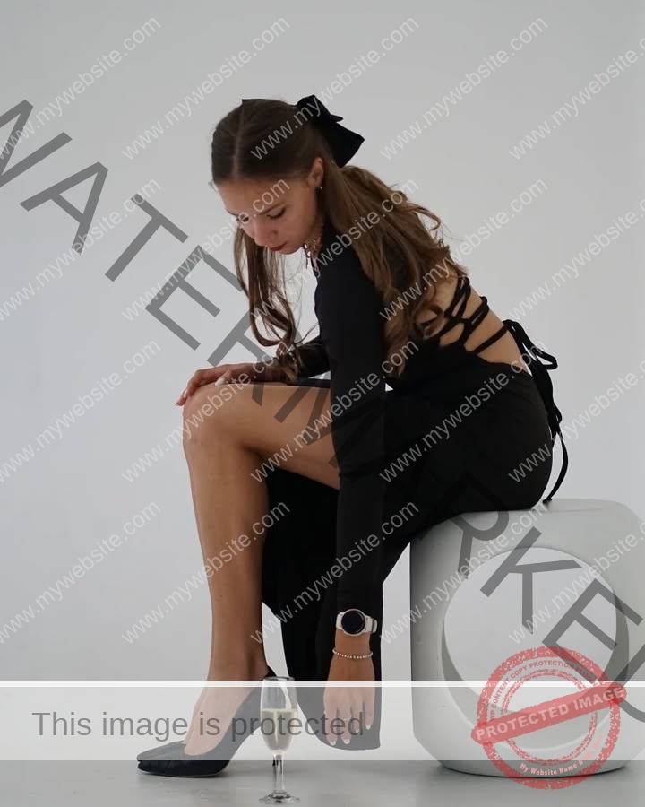 rufina-kotik-ukraine-rufina.kotik14-u18-02225 Rufina Kotik, track and field star from Ukraine, sits in a black dress on a white stool, leaning to touch her shoe by champagne.