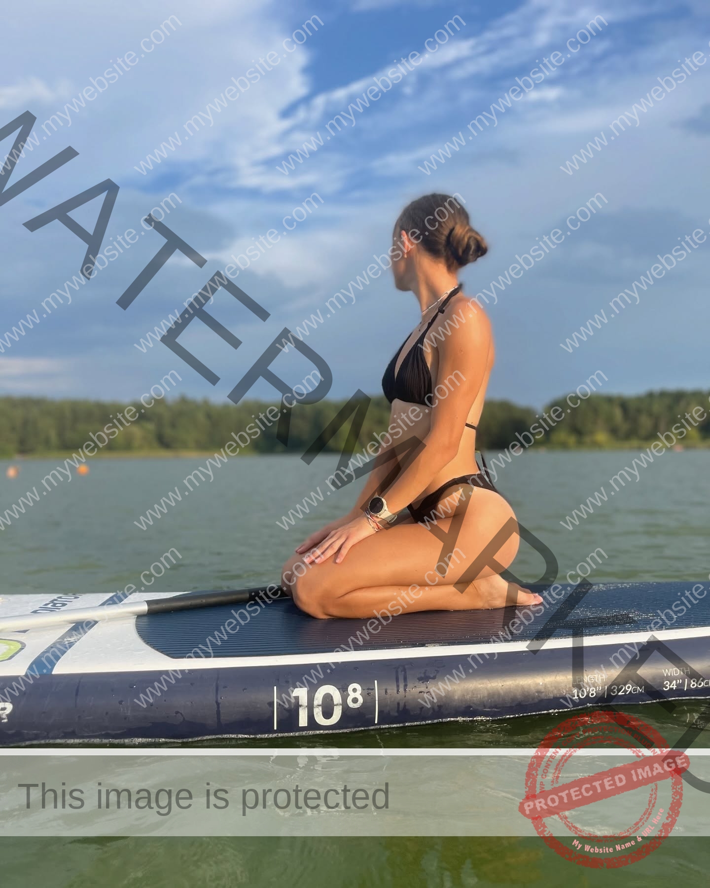 sasha-liubanska-ukraine-sasha_liubanska-u20-x-06042 Sasha Liubanska, track and field star from Ukraine, kneels on a paddleboard in a black bikini, gazing toward a tree-lined shore.