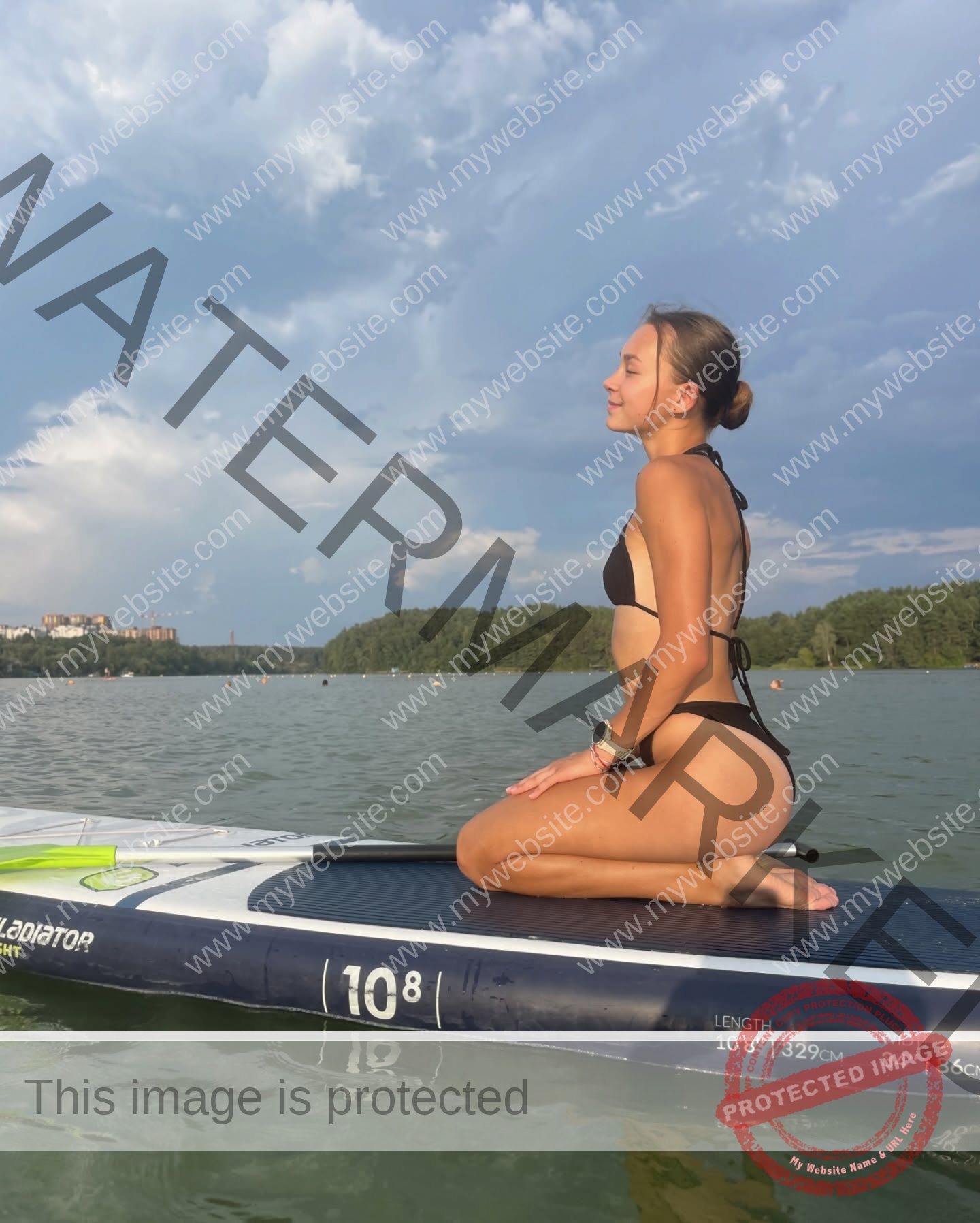 sasha-liubanska-ukraine-sasha_liubanska-u20-x-06043 Sasha Liubanska, track and field star from Ukraine, sits relaxed in a black bikini on a paddleboard on a calm lake with trees and buildings.