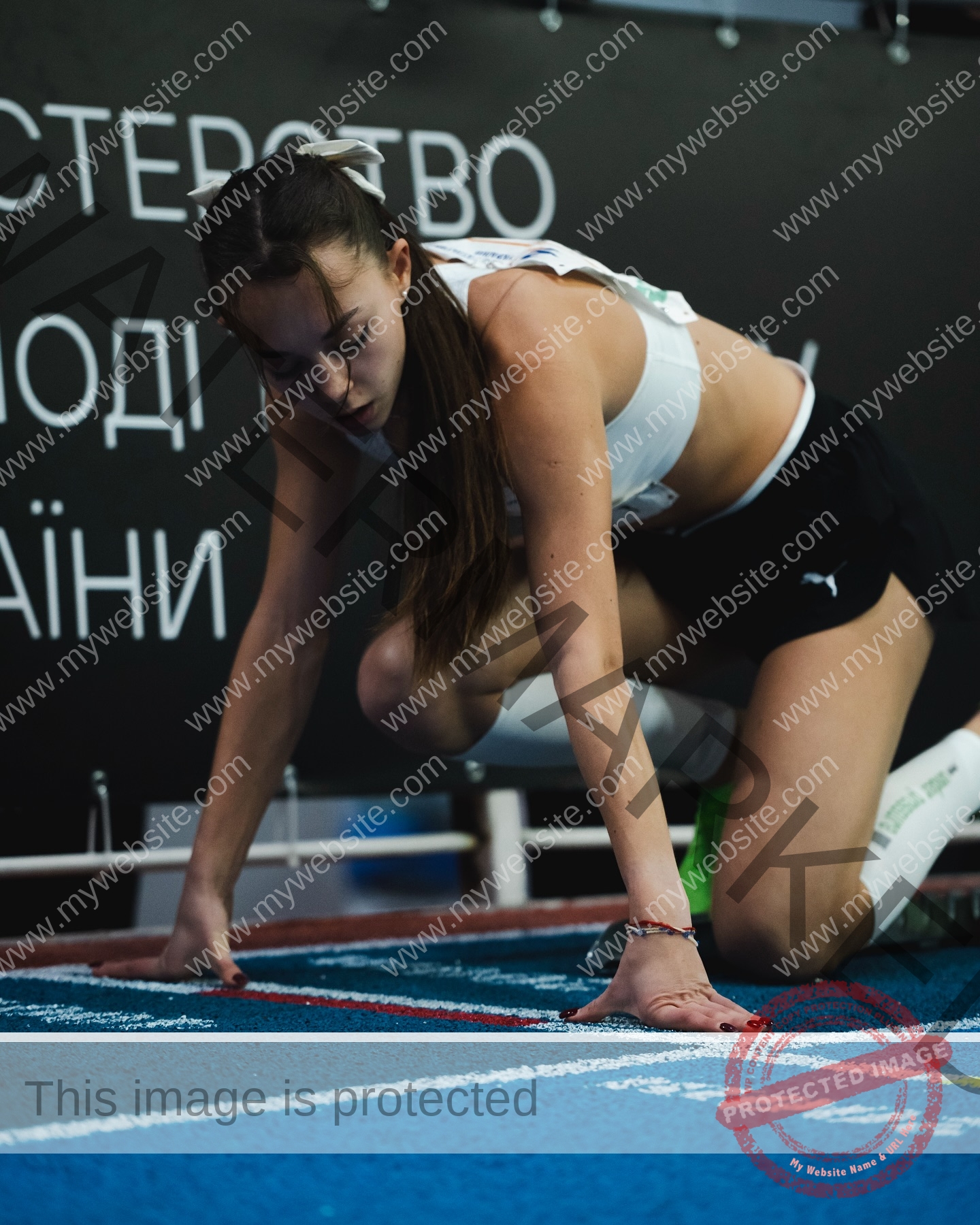 sasha-liubanska-ukraine-sasha_liubanska-u20-x-06051 Sasha Liubanska, track and field star from Ukraine, crouches at the starting line on a blue indoor track with Cyrillic writing behind.