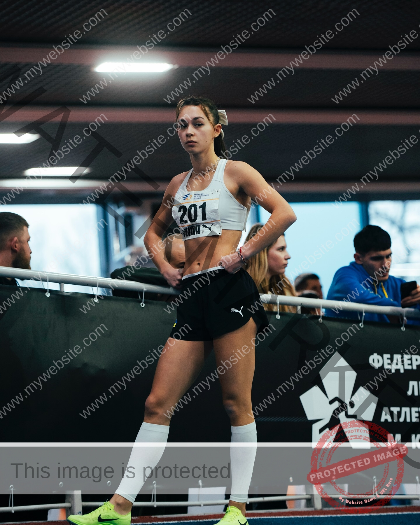 sasha-liubanska-ukraine-sasha_liubanska-u20-x-06052 Sasha Liubanska, track and field star from Ukraine, stands focused on an indoor track in sportswear, with people and a banner behind.