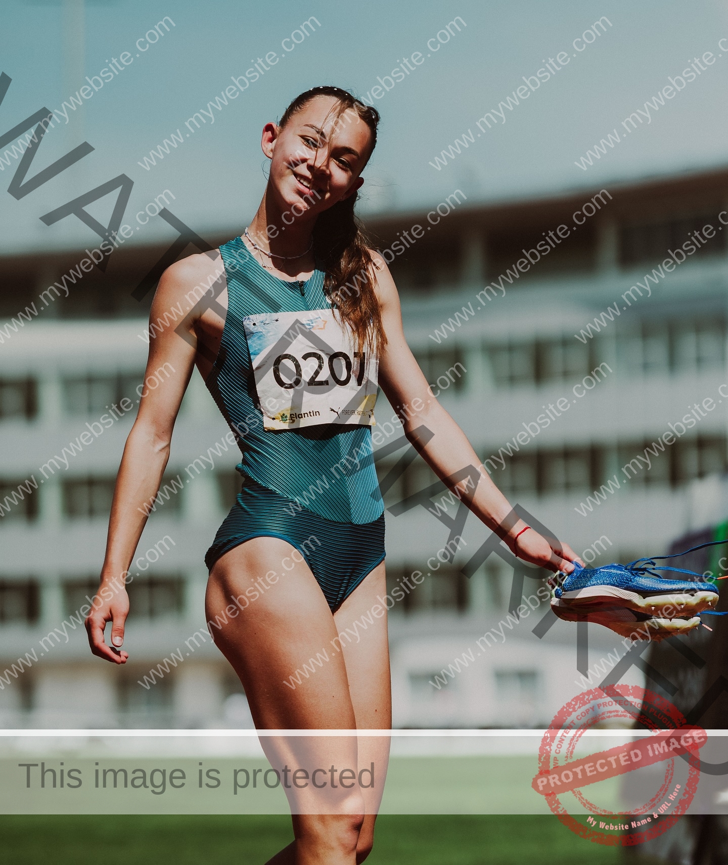 sasha-liubanska-ukraine-sasha_liubanska-u20-x-06058 Sasha Liubanska, track and field star from Ukraine, walks on the track in blue, smiling with her shoes in hand; stadium behind.