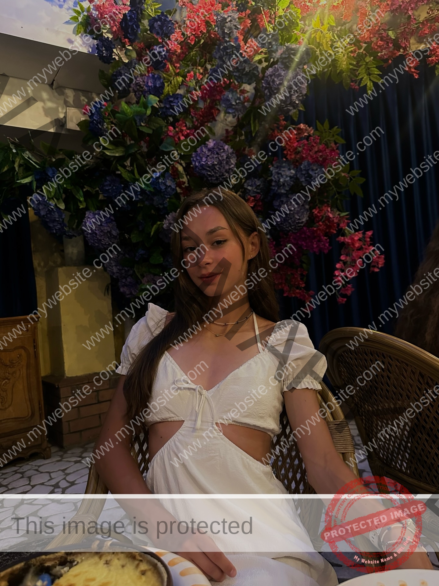 sasha-liubanska-ukraine-sasha_liubanska-u20-x-06060 Sasha Liubanska, track and field star from Ukraine, in a white dress at a wicker chair, smiles softly amid purple and pink flowers.