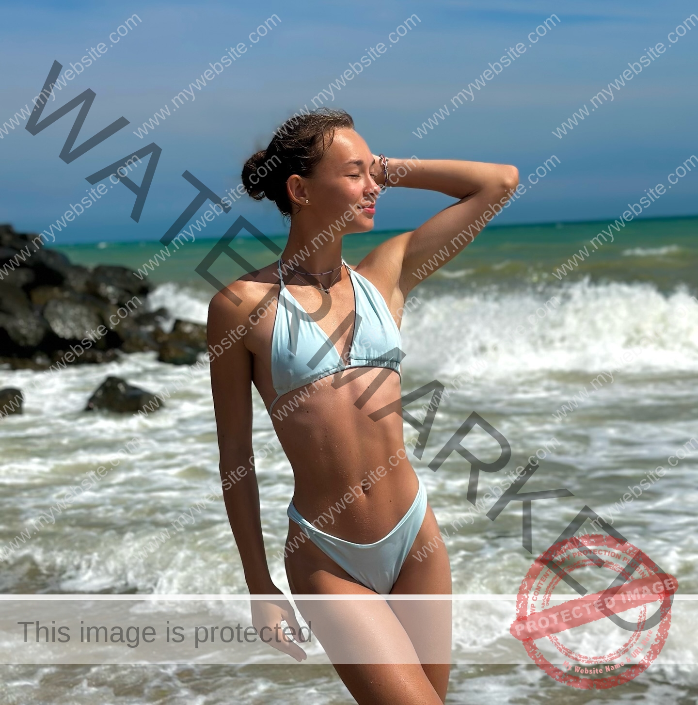 sasha-liubanska-ukraine-sasha_liubanska-u20-x-06063 Sasha Liubanska, track and field star from Ukraine, stands in a light blue bikini in shallow waves, smiling with one hand on her head.