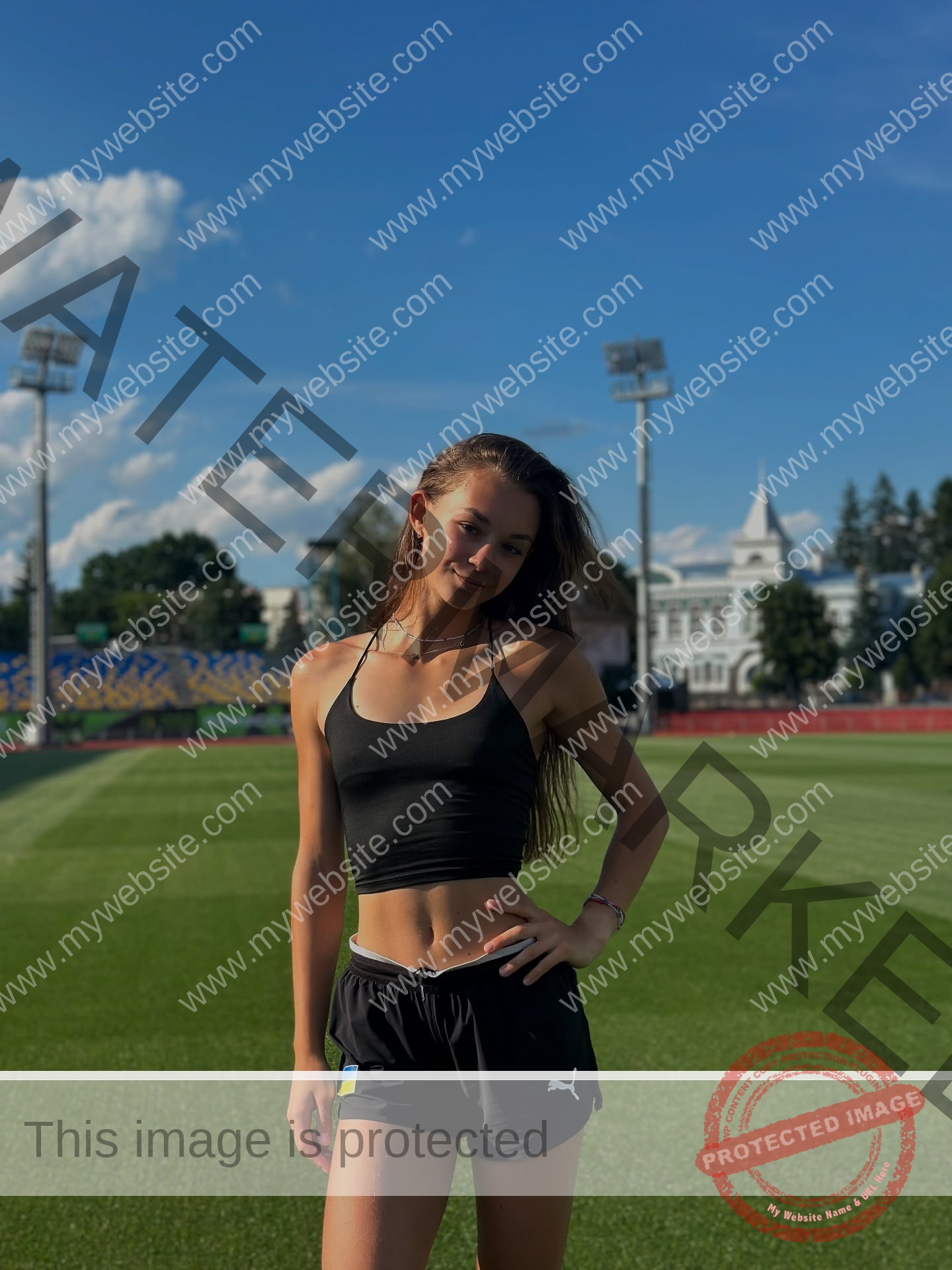 sasha-liubanska-ukraine-sasha_liubanska-u20-x-06065 Sasha Liubanska, track and field star from Ukraine, stands smiling on a green sports field under a blue sky with stadium lights behind.