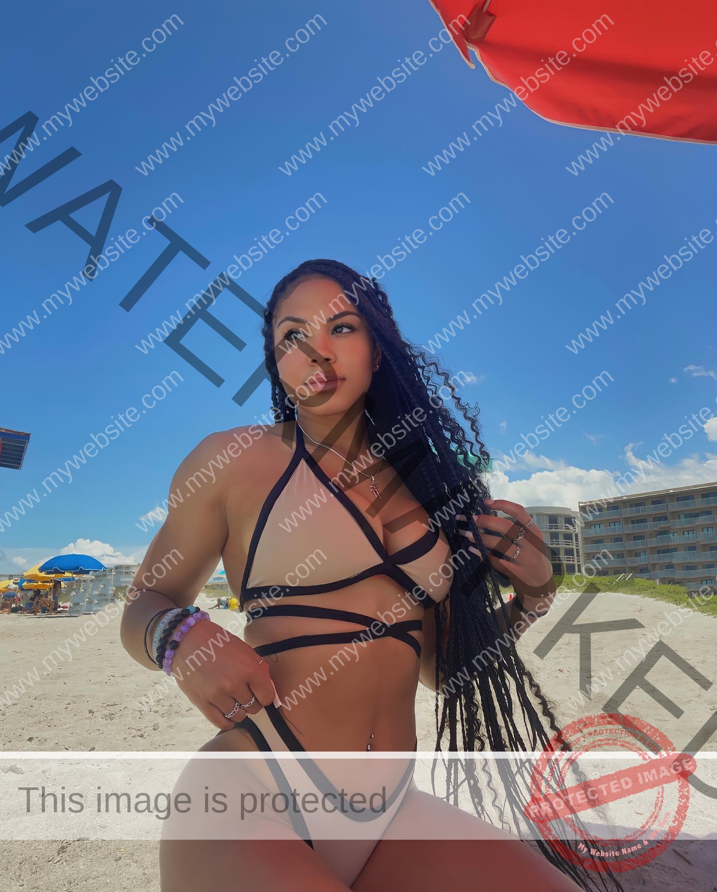 tatiana-aholou-canada-tatianaaholou-00402 Tatiana Ahoulou, track and field star from Canada, with long braids in a bikini poses on a sandy beach under a red umbrella.