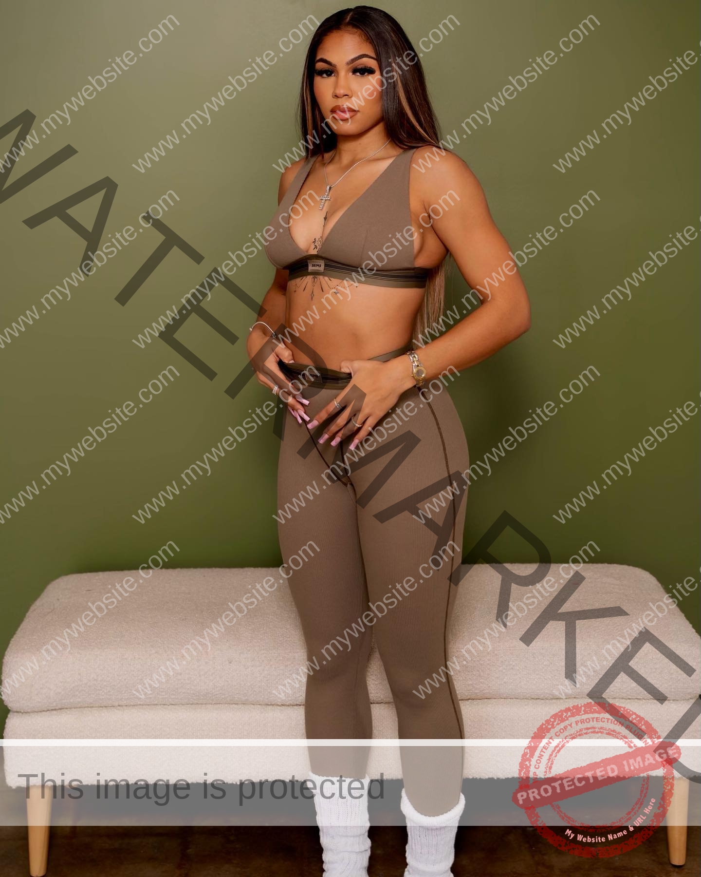 tatiana-aholou-canada-tatianaaholou-00431 Tatiana Ahoulou, track and field star from Canada, stands indoors in taupe activewear, hands on waist, by a beige bench.