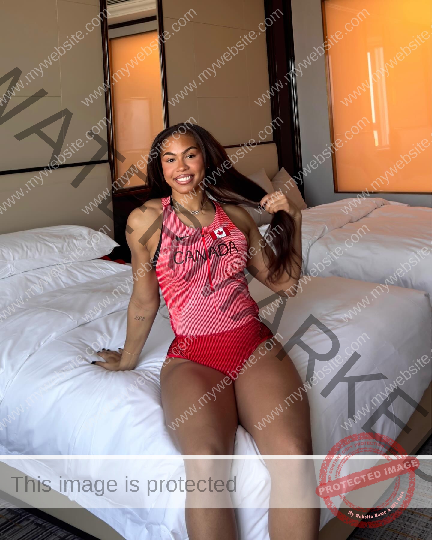tatiana-aholou-canada-tatianaaholou-00485 Tatiana Ahoulou, track and field star from Canada, sits on a hotel bed in a red uniform, smiling and holding her long hair.