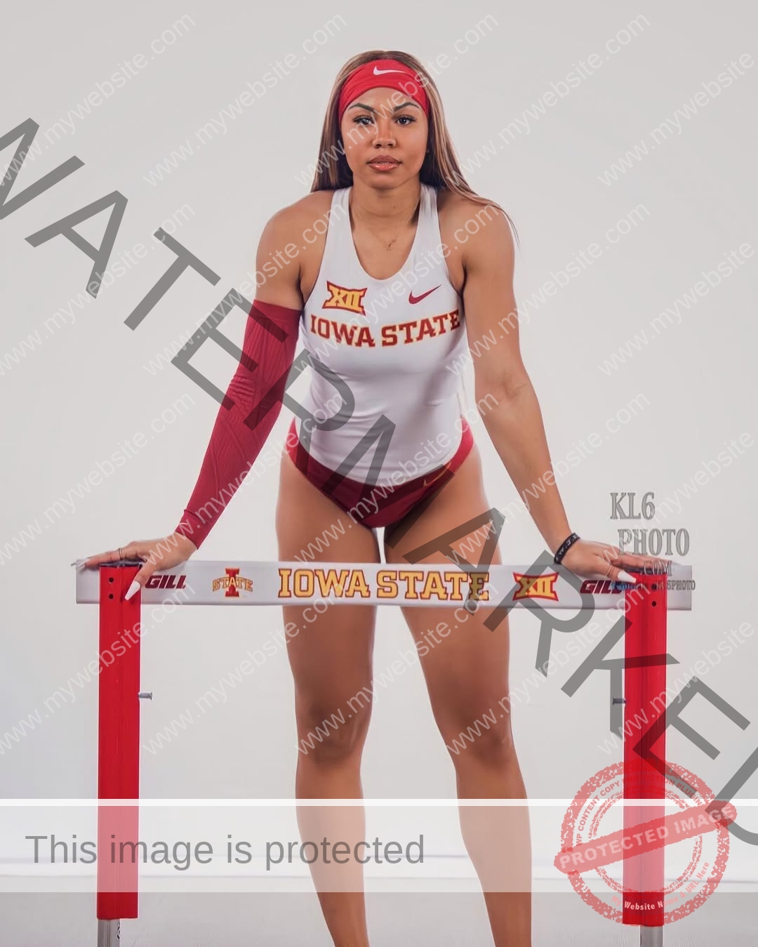 tatiana-aholou-canada-tatianaaholou-00510 Tatiana Ahoulou, track and field star from Canada, stands in an Iowa State uniform behind a hurdle with a red headband and arm sleeve.