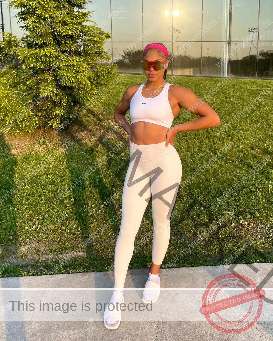 tatiana-aholou-canada-tatianaaholou-00527 Tatiana Ahoulou, track and field star from Canada, poses hands-on-hips in white sportswear on a sidewalk near a glass building.