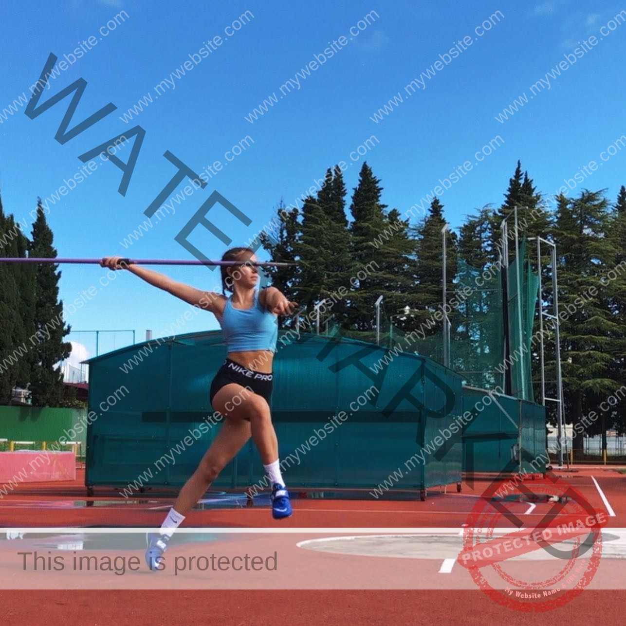viktoriya-antipova-russia-viichkaaaaa-03465 Viktoriya Antipova, track and field star from Russia, strides on a red track, arm back with javelin; trees visible behind her.