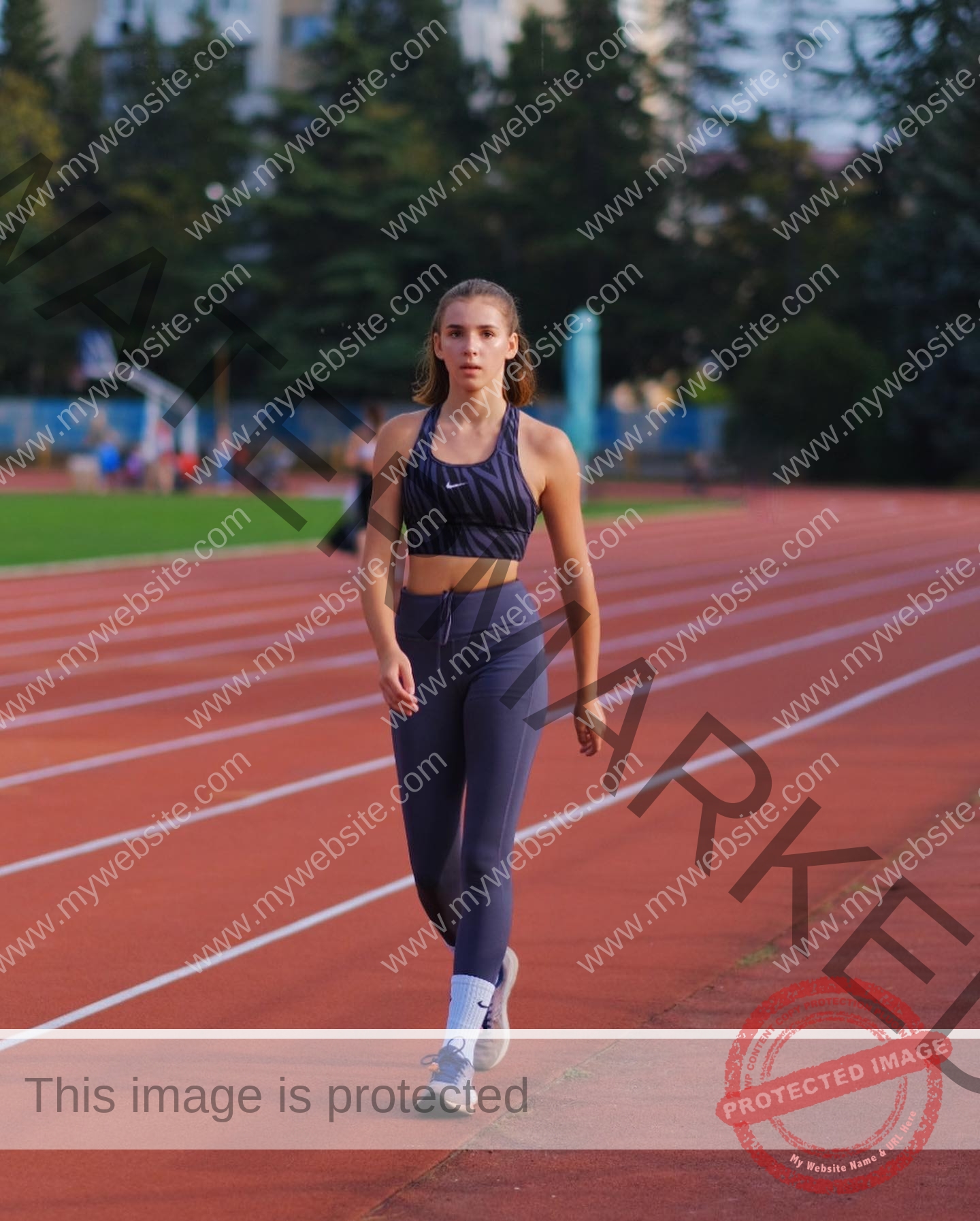viktoriya-antipova-russia-viichkaaaaa-03469 Viktoriya Antipova, track and field star from Russia, walks on a red running track outdoors, focused amid green trees.