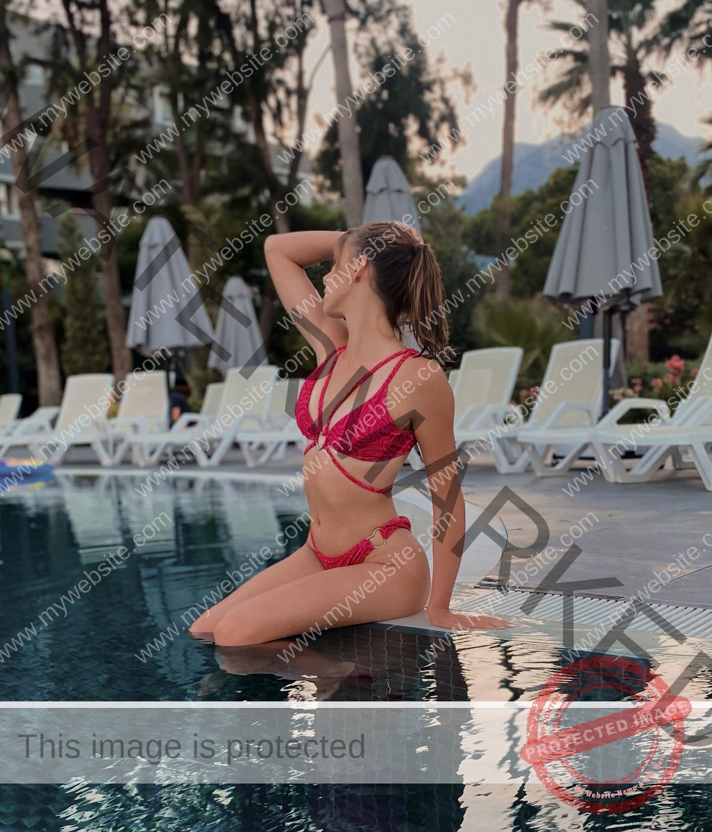 viktoriya-antipova-russia-viichkaaaaa-03505 Viktoriya Antipova, track and field star from Russia, kneels by a pool in a pink bikini with one hand in her hair; poolside chairs behind.