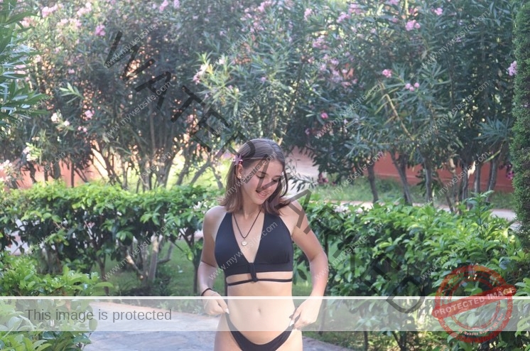 viktoriya-antipova-russia-viichkaaaaa-03522 Viktoriya Antipova, track and field star from Russia, wears a black bikini outdoors, smiling with a flower in her hair by green bushes.