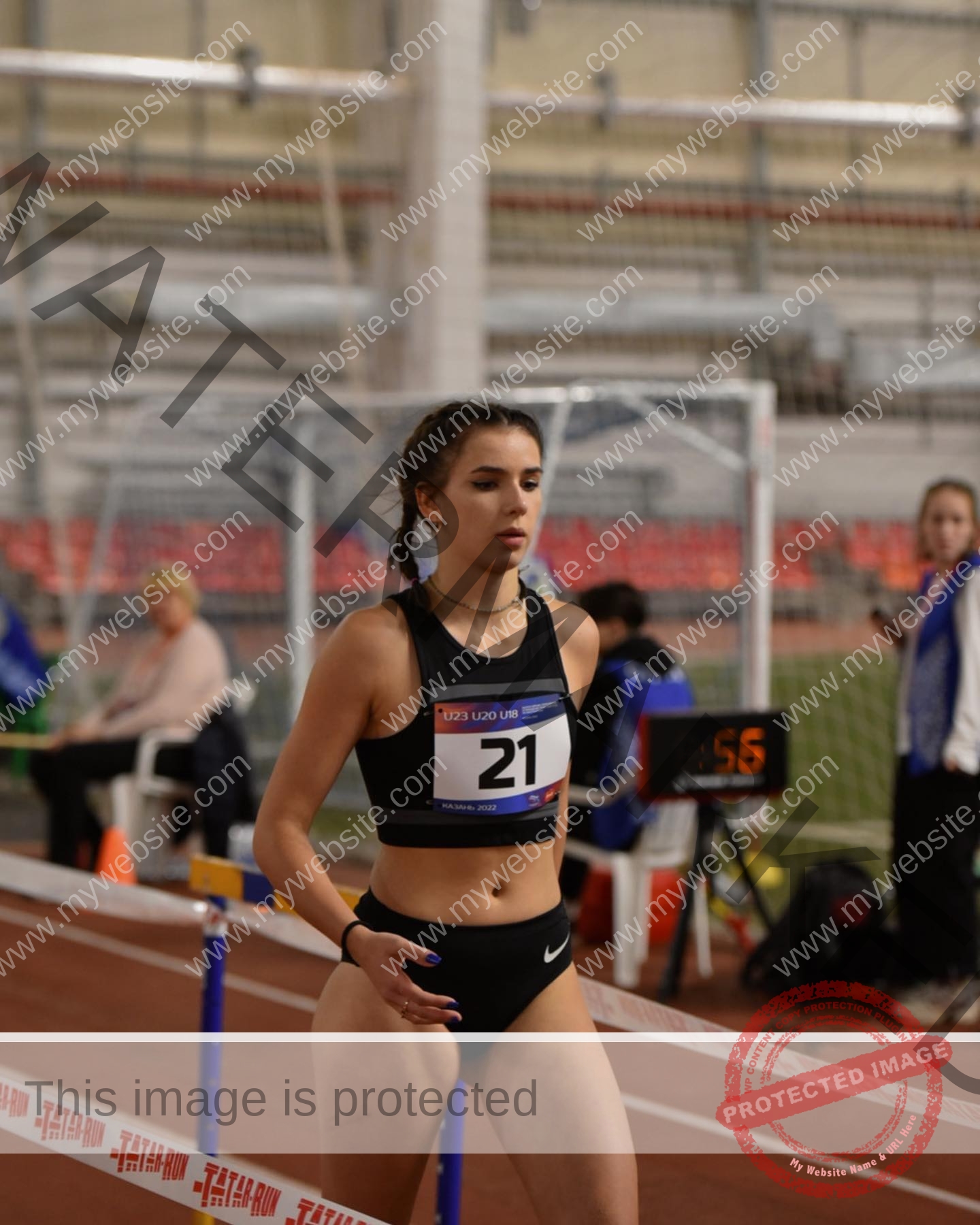 viktoriya-antipova-russia-viichkaaaaa-03538 Viktoriya Antipova, track and field star from Russia, wears black sportswear and bib 21 as she walks past hurdles on an indoor track.