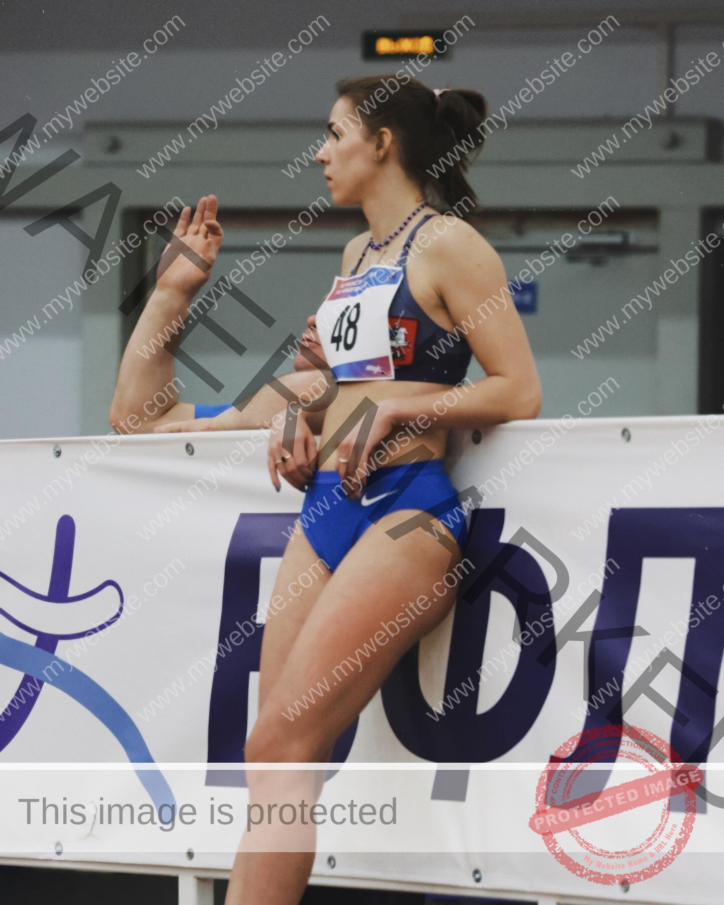 viktoriya-antipova-russia-viichkaaaaa-03626 Viktoriya Antipova, track and field star from Russia, in blue, #48, leans on a barrier at an indoor track with Russian text.