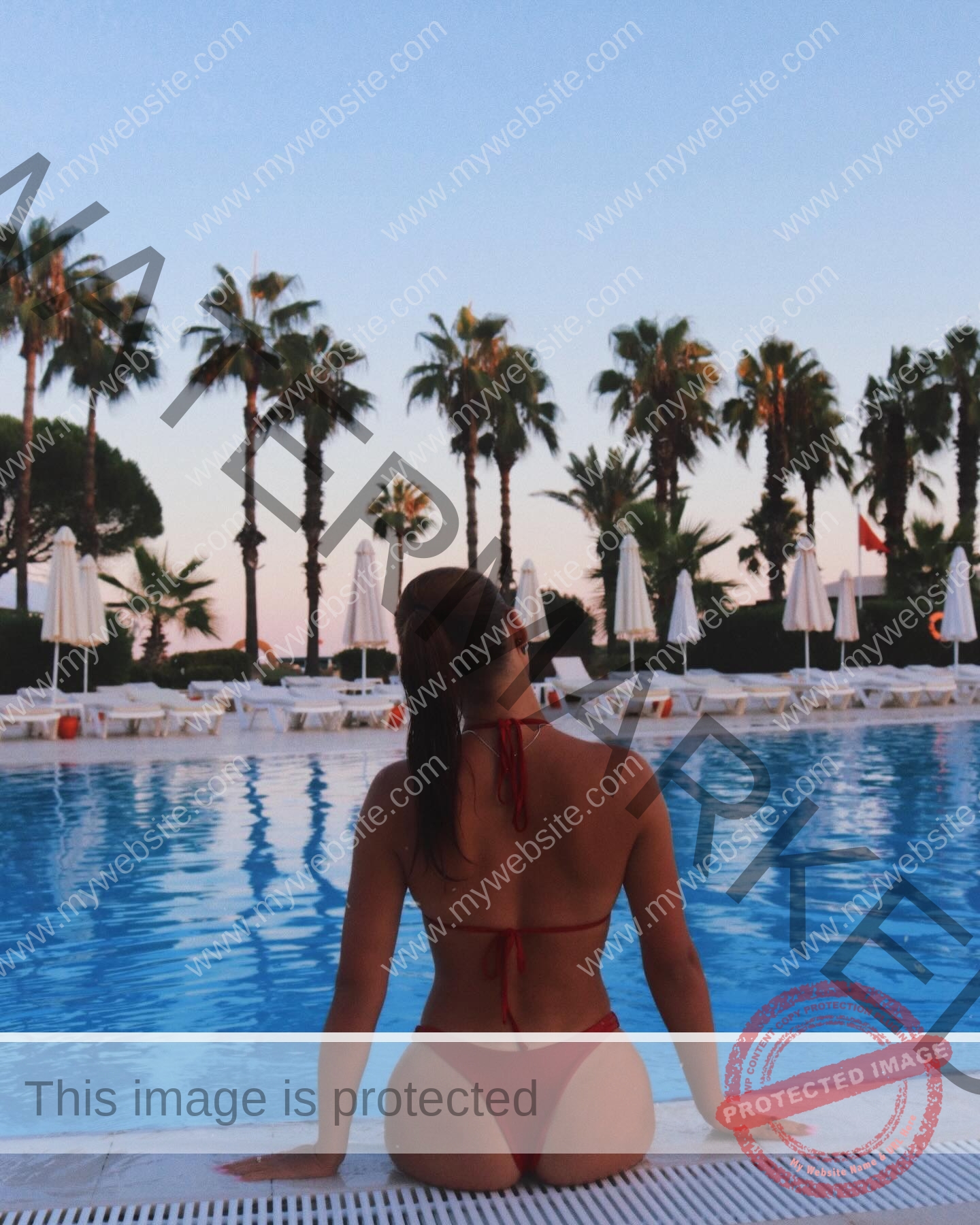 viktoriya-antipova-russia-viichkaaaaa-03670 Viktoriya Antapova, track and field star from Russia, in a red bikini sits at a pool's edge, sunset sky and palms behind her.