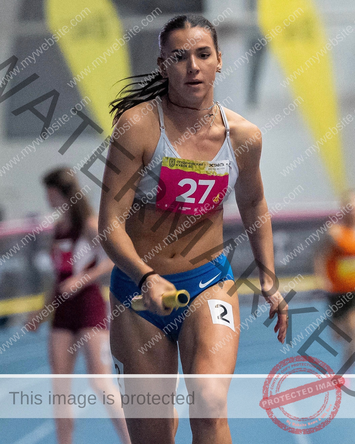 viktoriya-antipova-russia-viichkaaaaa-03685 Viktoriya Antapova, track and field star from Russia, runs in blue shorts and white top with baton; bib number 27A. Other runners behind.