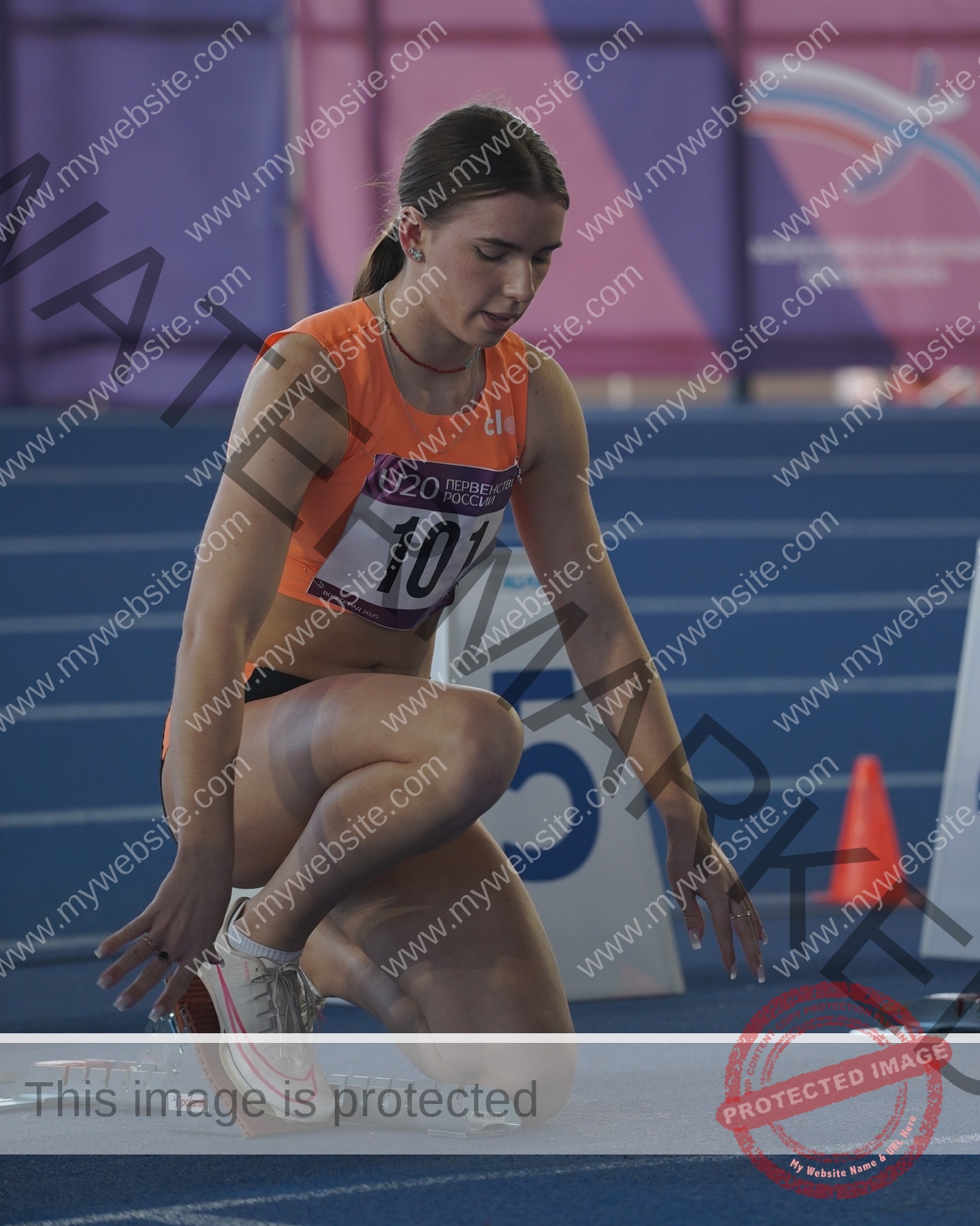 viktoriya-antipova-russia-viichkaaaaa-03692 Viktoriya Antipova, track and field star from Russia, in an orange top kneels at a blue indoor starting block, race bib 101 on.
