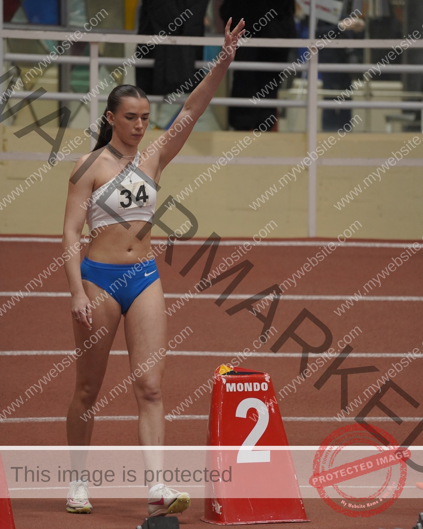 viktoriya-antipova-russia-viichkaaaaa-03713 Viktoriya Antipova, track and field star from Russia, wears a white crop top 34 and blue shorts, raising one arm by block 2 indoors.