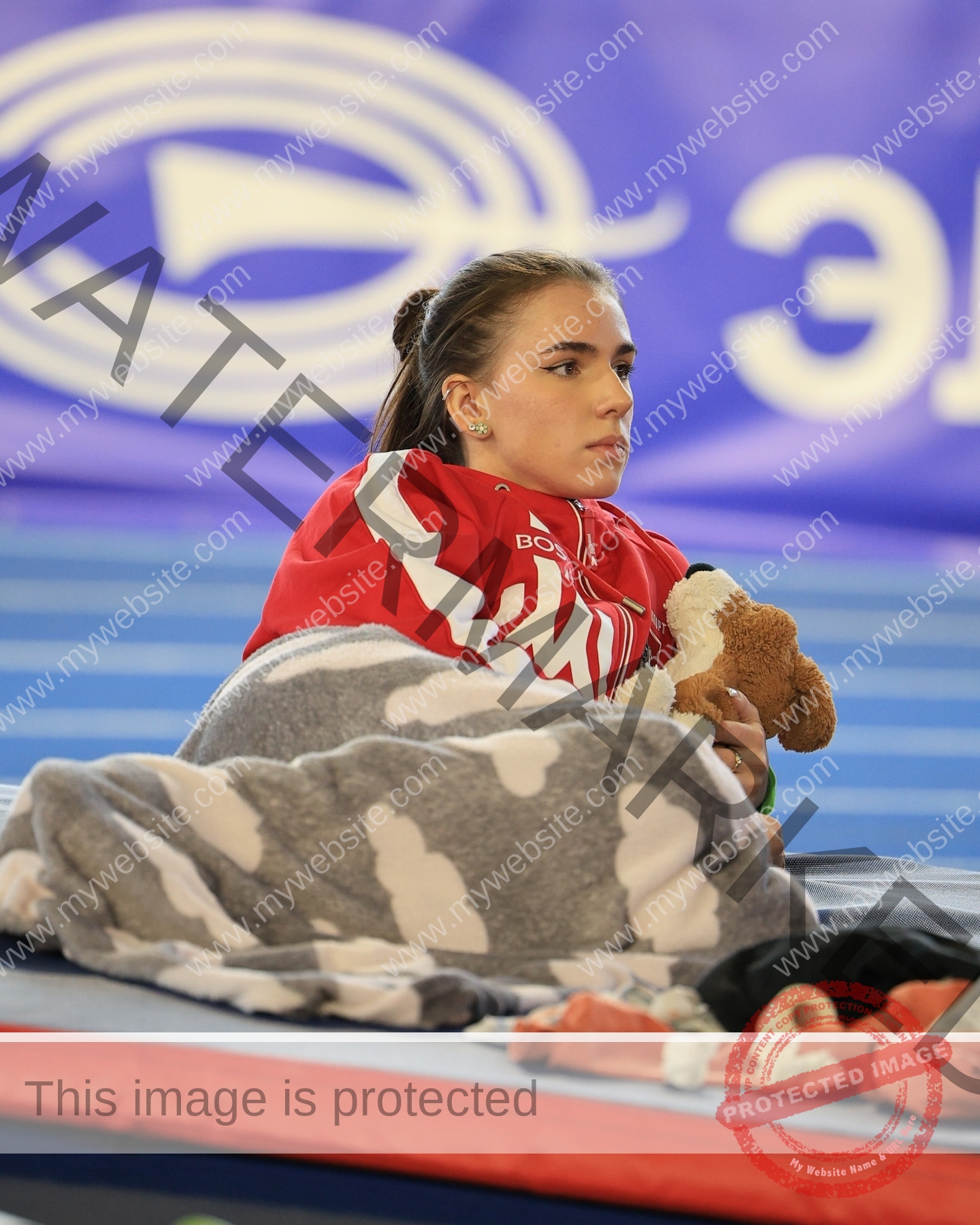 viktoriya-antipova-russia-viichkaaaaa-03717 Viktoriya Antipova, track and field star from Russia, sits on a mat in a red jacket, wrapped in a cloud-pattern blanket, holding a stuffed dog toy.