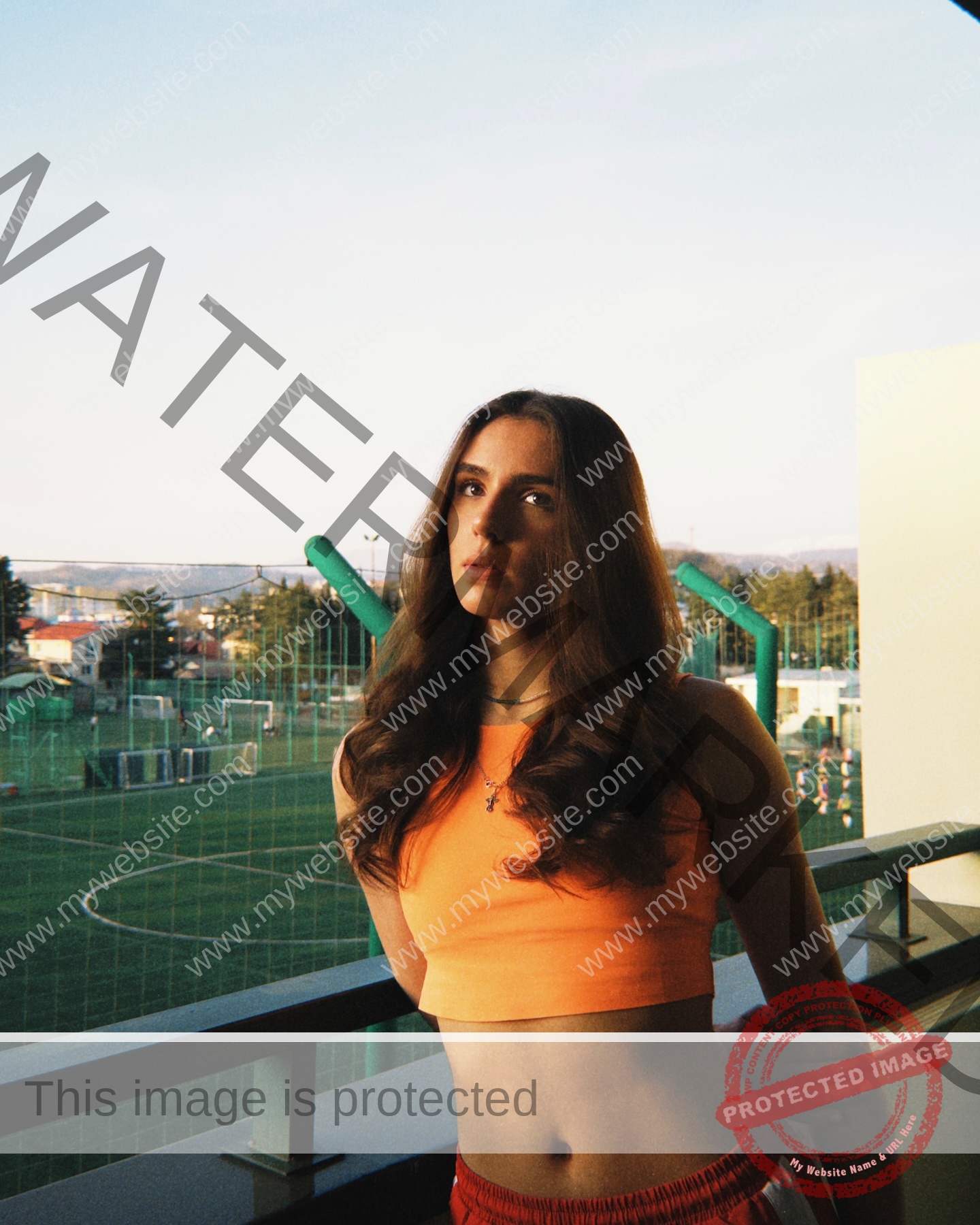 viktoriya-antipova-russia-viichkaaaaa-03720 Viktoriya Antapova, track and field star from Russia, with long wavy hair in orange crop top and red shorts on a balcony at sunset.