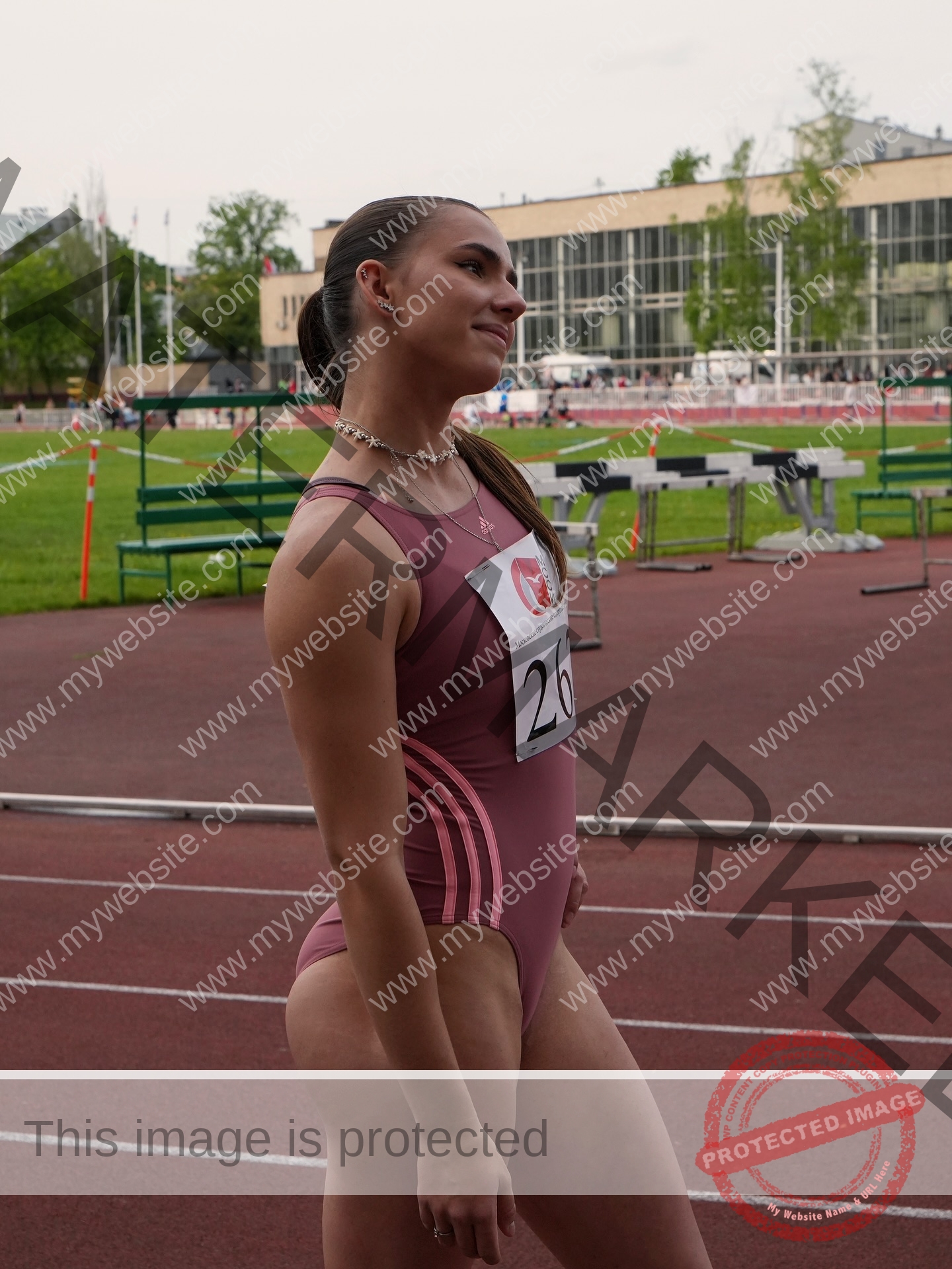 viktoriya-antipova-russia-viichkaaaaa-03731 Viktoriya Antipova, track and field star from Russia, wears a mauve bodysuit and bib 26, smiling on the track with bleachers behind.