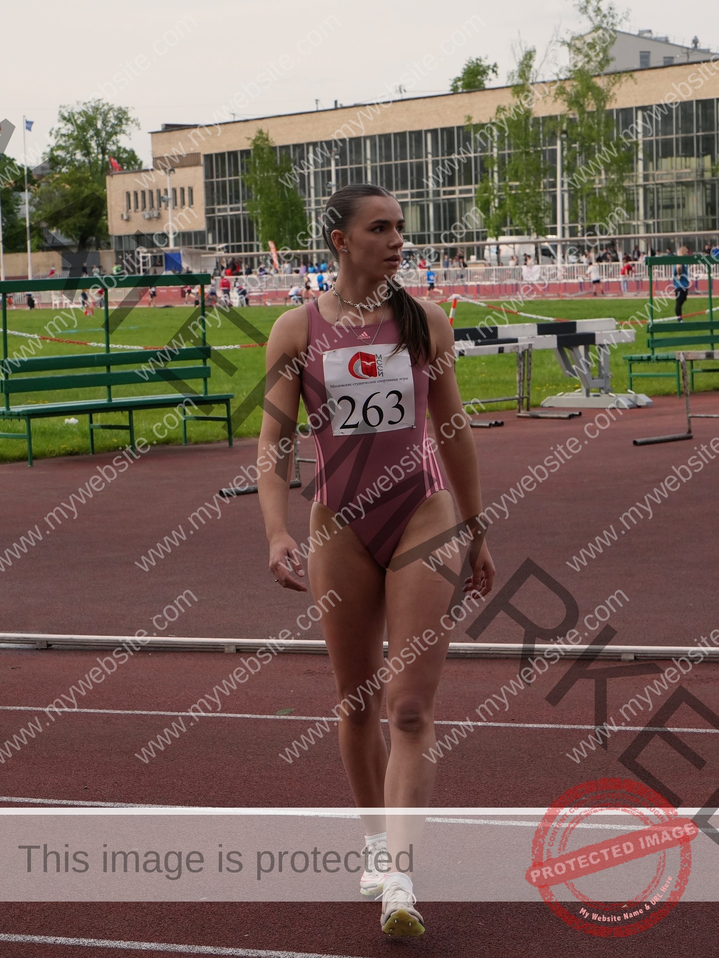 viktoriya-antipova-russia-viichkaaaaa-03732 Viktoriya Antipova, track and field star from Russia, in a pink bodysuit (#263) walks on an outdoor running track; benches behind.