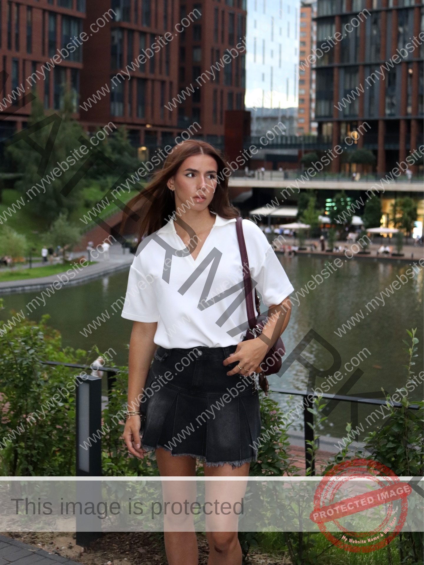 viktoriya-antipova-russia-viichkaaaaa-03741 Viktoriya Antapova, track and field star from Russia, stands near a canal in a white blouse and black skirt amid modern buildings.