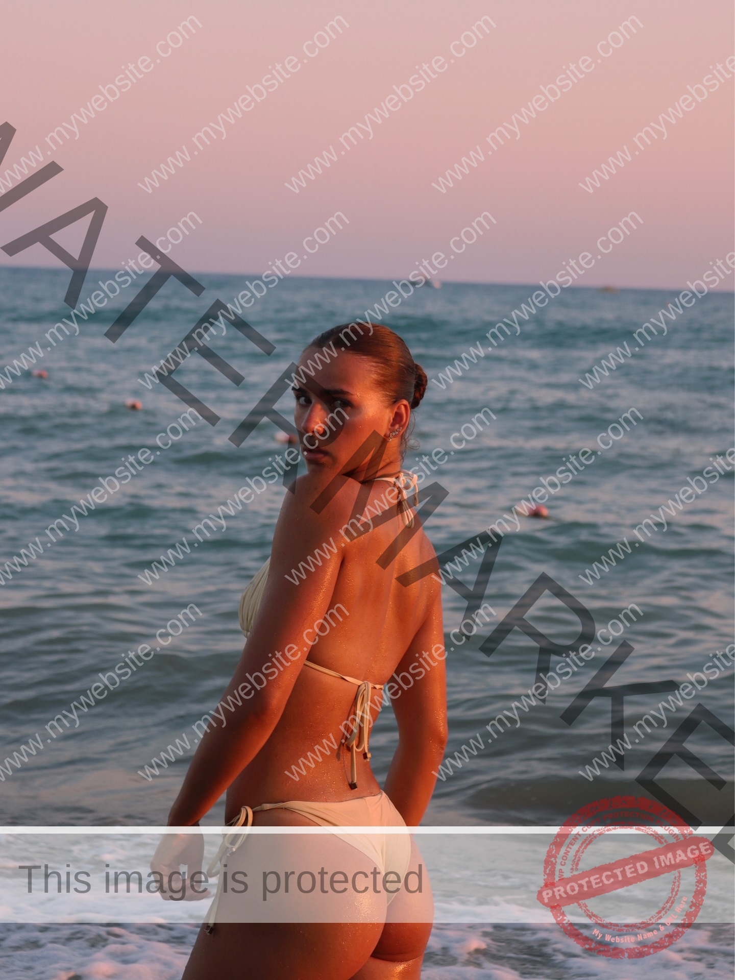 viktoriya-antipova-russia-viichkaaaaa-03771 Viktoriya Antapova, track and field star from Russia, in a light bikini on the shore looks back at the camera; ocean and pastel sunset behind.