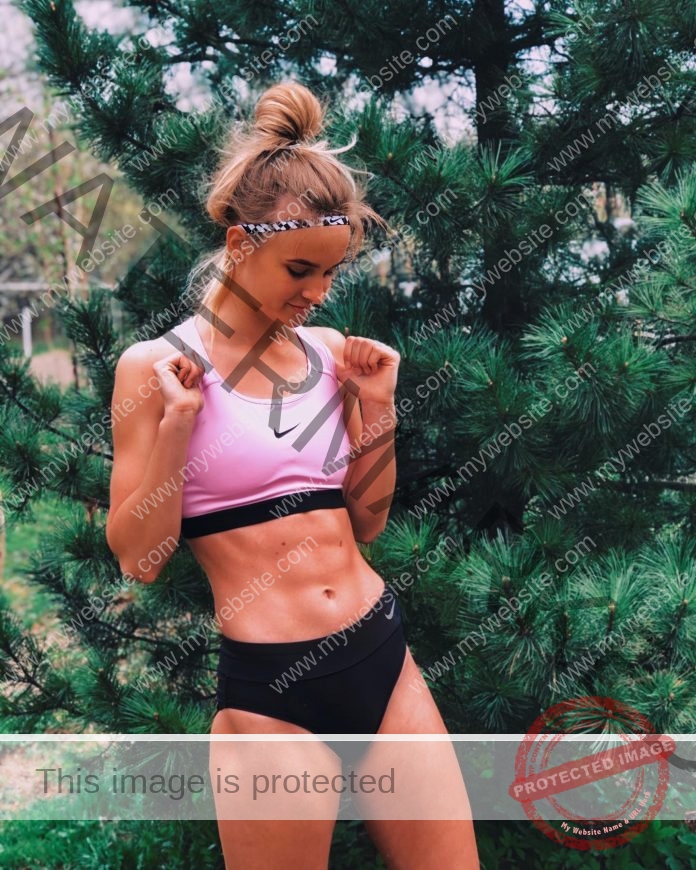 Yana Zaytseva, track and field star from Russia, wears a pink sports bra and black shorts outdoors by a pine tree, looking down.