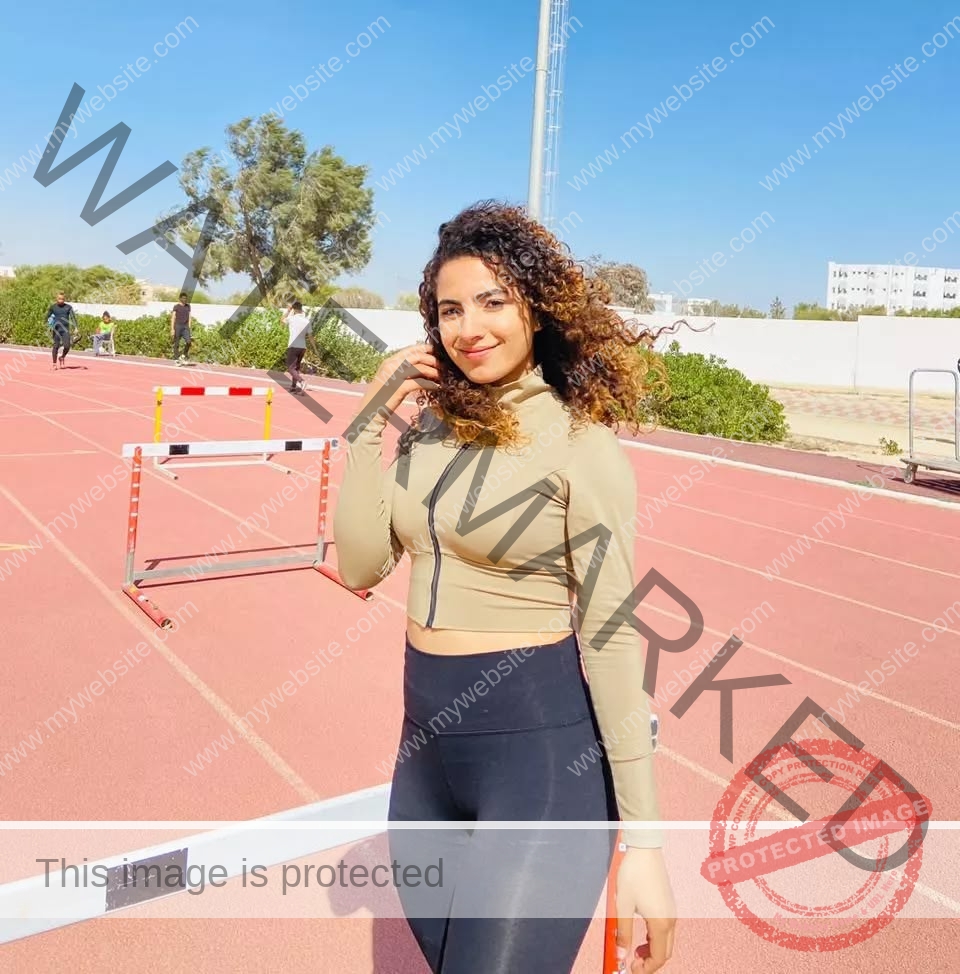 yassmine-baccar-tunisia-yassmine_baccar-08432 Yassmire Baccar, track and field star from Tunisia, smiles on a sunlit track in tan top and black leggings by hurdles and greenery.