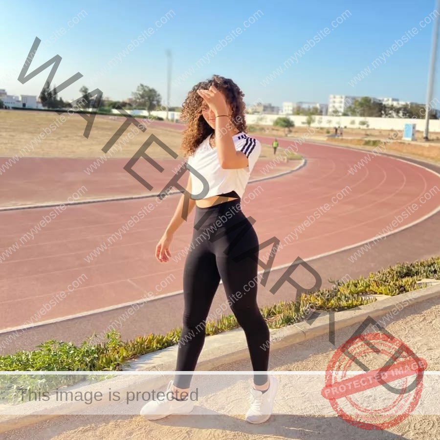 yassmine-baccar-tunisia-yassmine_baccar-08433 Yassmire Baccar, track and field star from Tunisia, stands by an outdoor track in a white crop top and black leggings, shading her eyes.