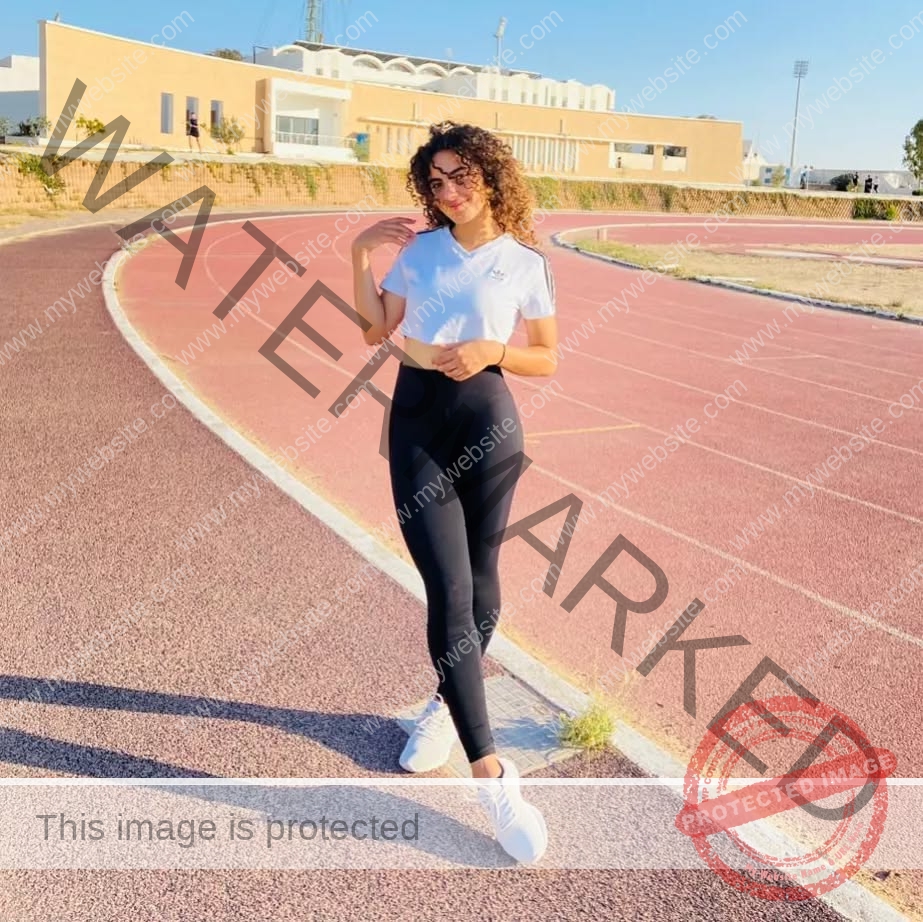yassmine-baccar-tunisia-yassmine_baccar-08434 Yassmire Baccar, track and field star from Tunisia, in a white crop top and black leggings smiles on a red track; building behind.