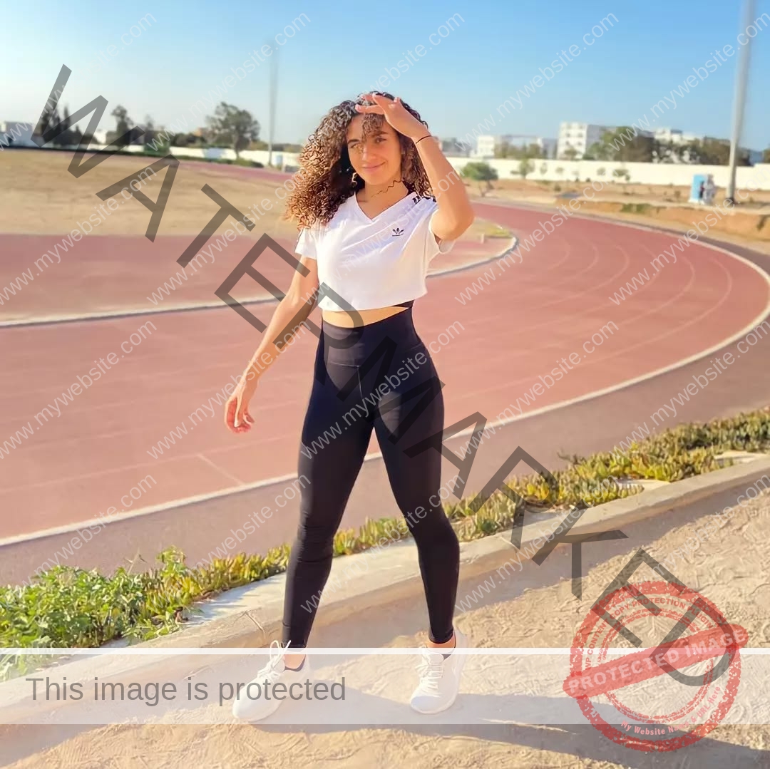 yassmine-baccar-tunisia-yassmine_baccar-08435 Yassmire Baccar, track and field star from Tunisia, stands by a sunny running track, touching her hair in a white crop top and leggings.