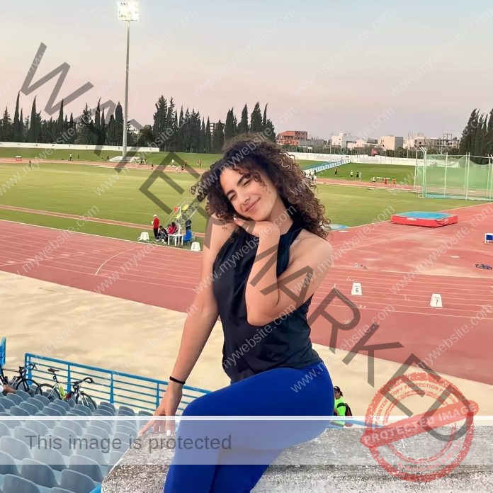Yassmire Baccar, track and field star from Tunisia, with curly hair, sits in a stadium at sunset by a track, hand under chin.