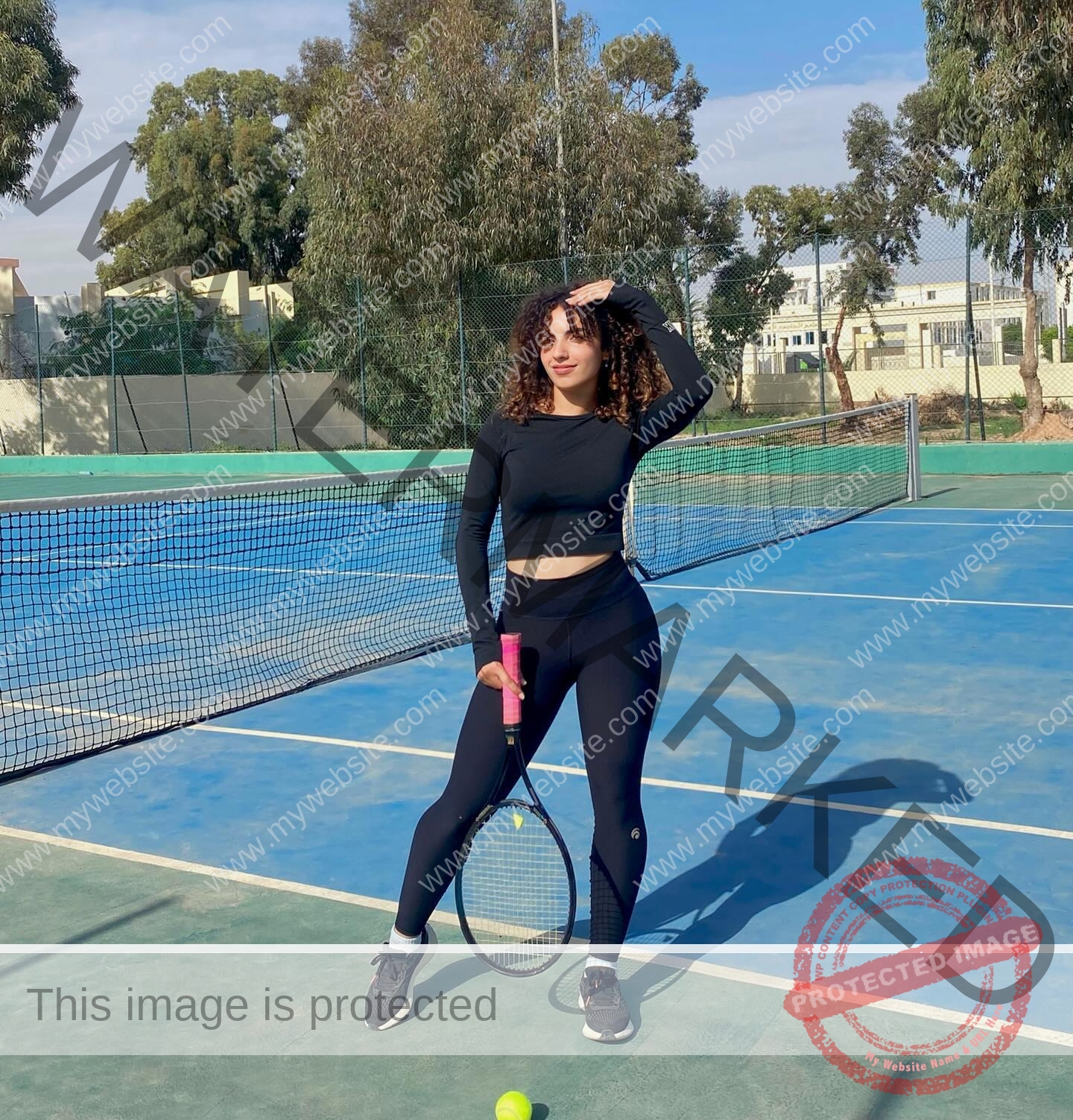 yassmine-baccar-tunisia-yassmine_baccar-08463 Yassmire Baccar, track and field star from Tunisia, in black athletic wear on a blue court, holds a pink-grip racket confidently.