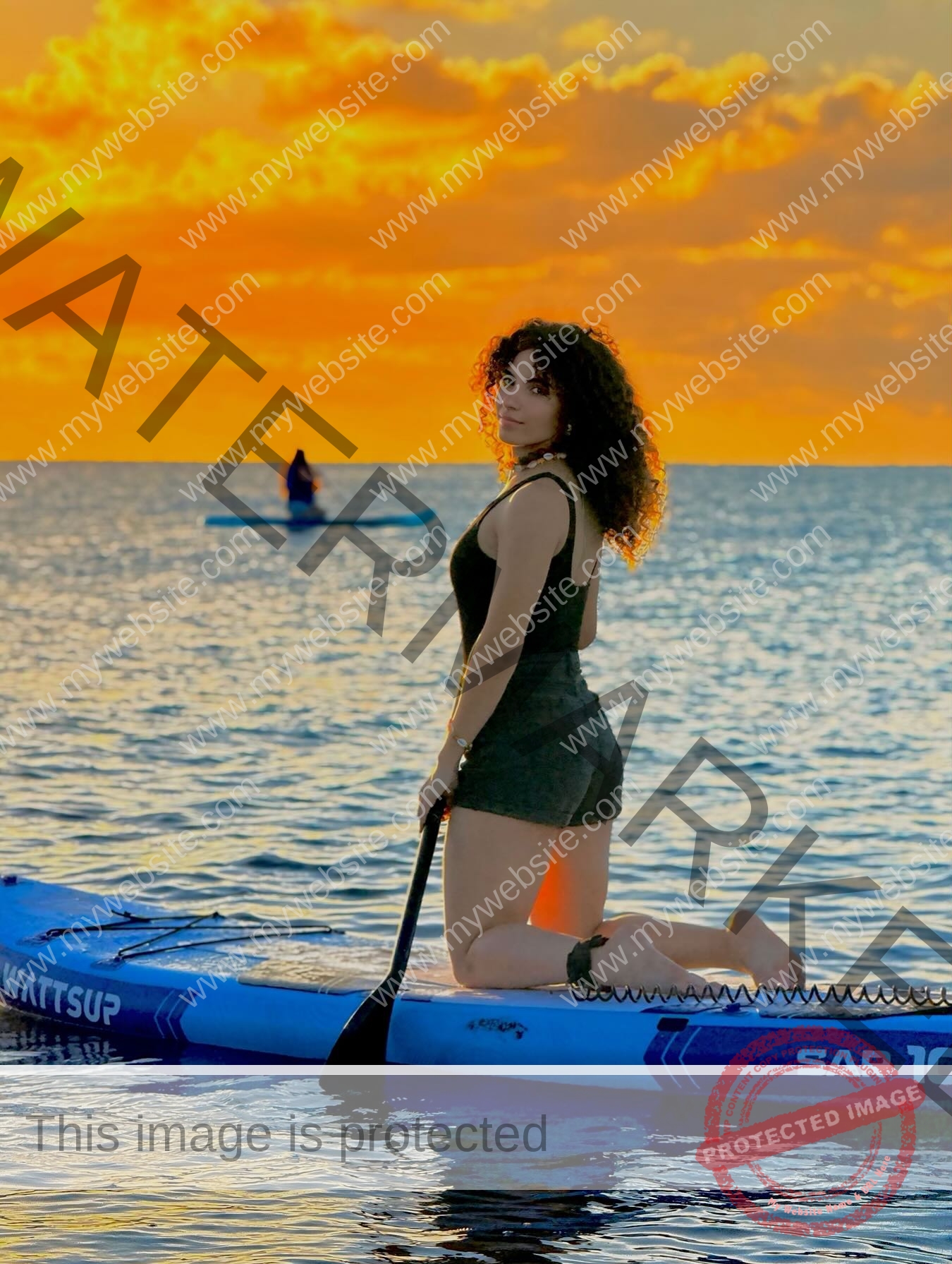 yassmine-baccar-tunisia-yassmine_baccar-08496 Yassmire Baccar, track and field star from Tunisia, kneels on a blue paddleboard at sunset with vivid orange sky and calm water.