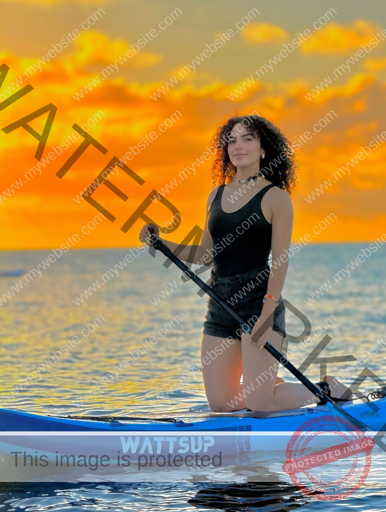 yassmine-baccar-tunisia-yassmine_baccar-08497 Yassmire Baccar, track and field star from Tunisia, with curly hair, kneels on a blue WATTSUP paddleboard at sunset.