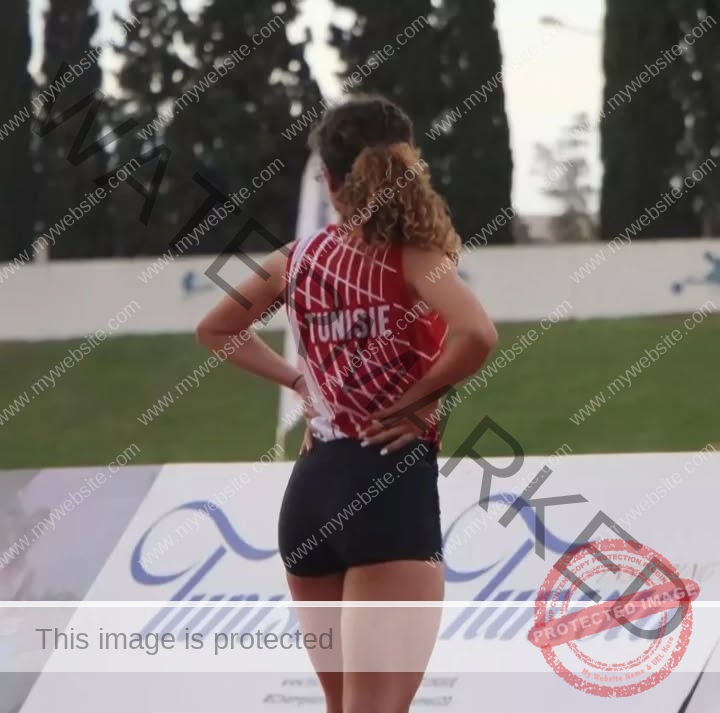 yassmine_baccartagged-08498 Yassmire Baccar, track and field star from Tunisia, stands on an outdoor track in a red and white Tunisie jersey, hands on hips.