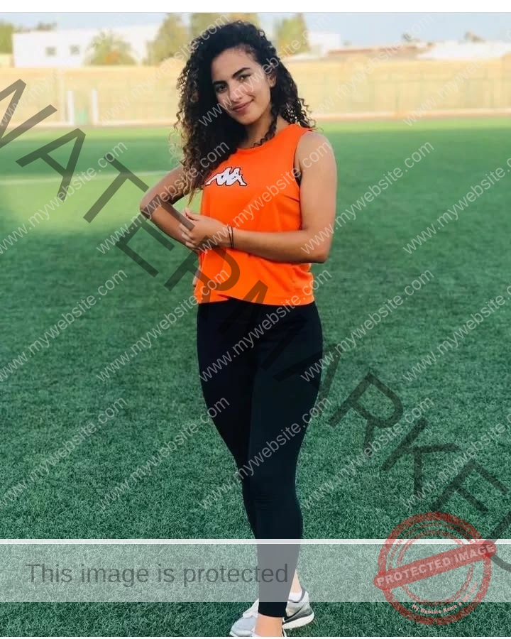yassmine_baccartagged-08502 Yassmire Baccar, track and field star from Tunisia, stands on a green sports field, smiling in orange top and black leggings.