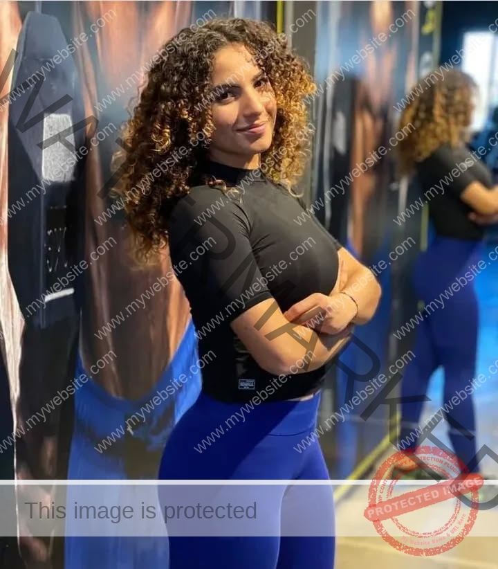 yassmine_baccartagged-08503 Yassmire Baccar, track and field star from Tunisia, with curly hair, in black top and blue leggings, stands smiling in a gym.