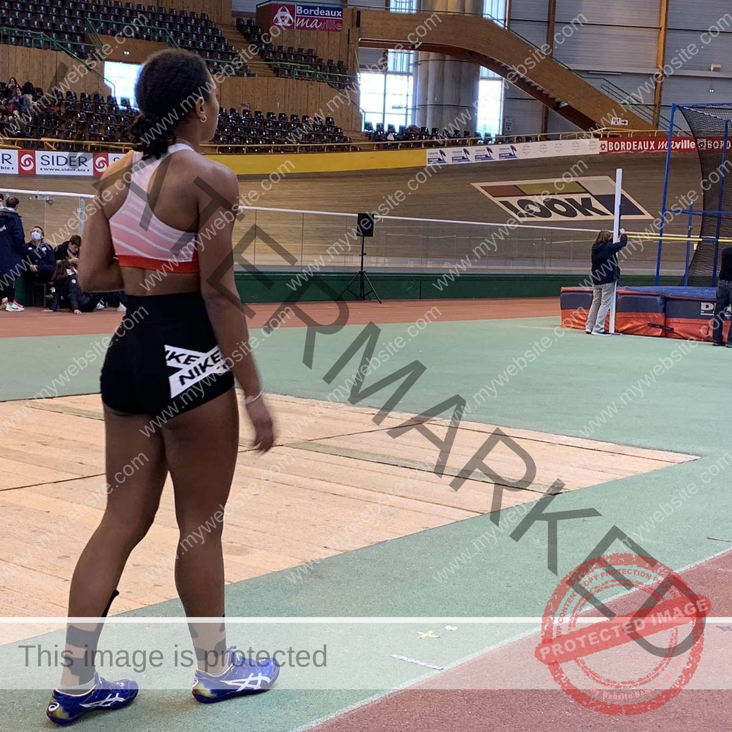 zola-ndouma-mona-france-zolaathle-09268 Zola Ndouma Mona, track and field star from France stands on an indoor track, sports bra, shorts, tape on leg; official with pole behind.