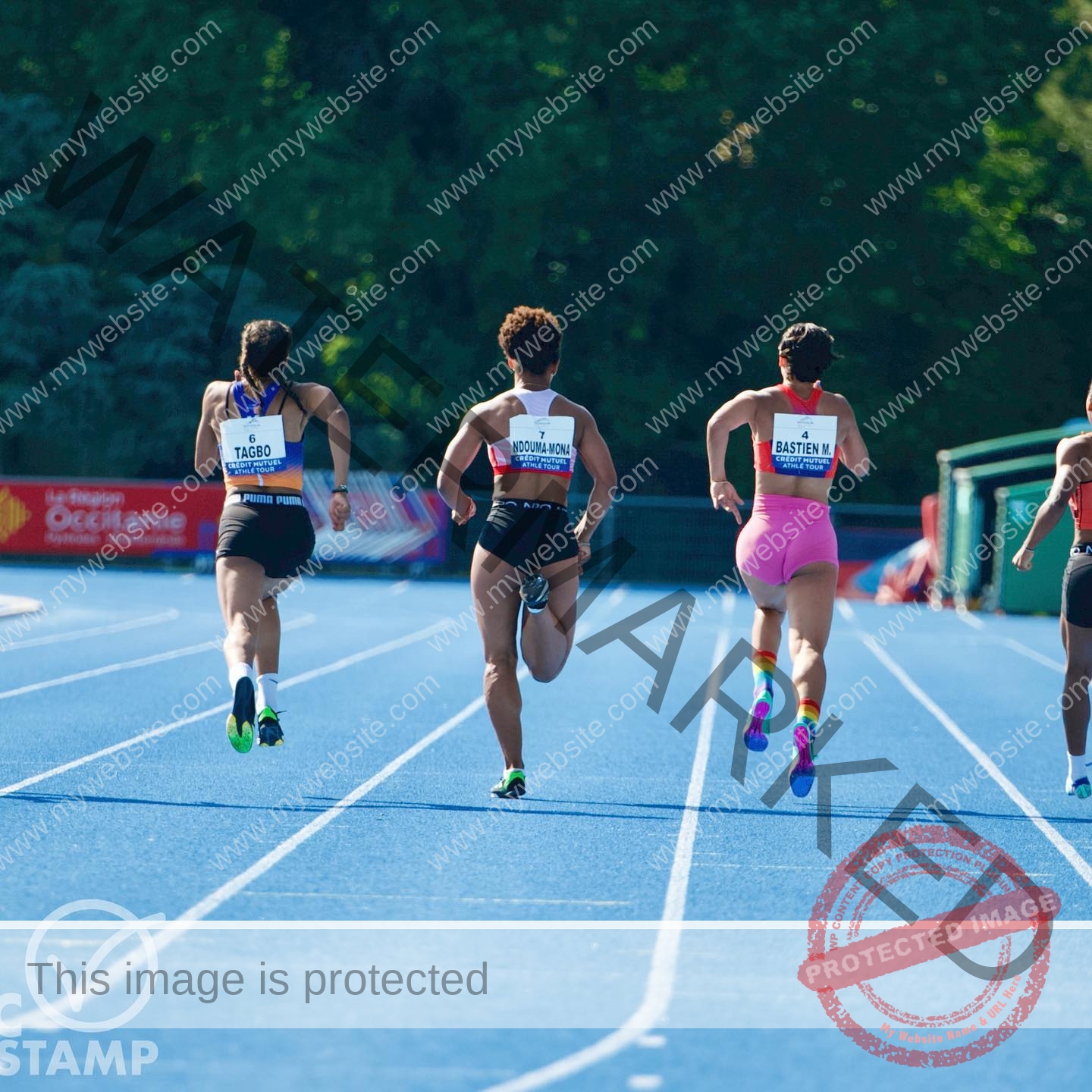zola-ndouma-mona-france-zolaathle-09288 Zola Ndouma Mona, track and field star from France sprints on a blue outdoor track among three others, with trees and a stadium banner.
