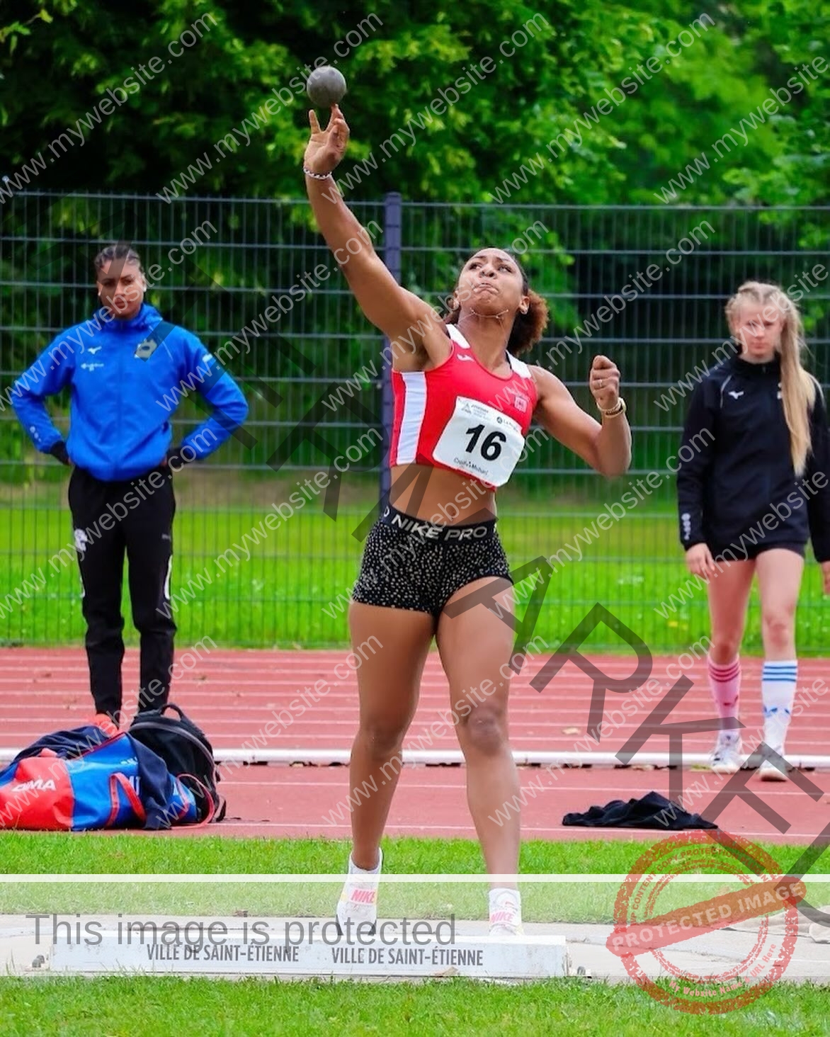 zola-ndouma-mona-france-zolaathle-09319 Zola Ndouma Mona, track and field star from France, in red and white jersey throws shot put as two others watch on the outdoor track.
