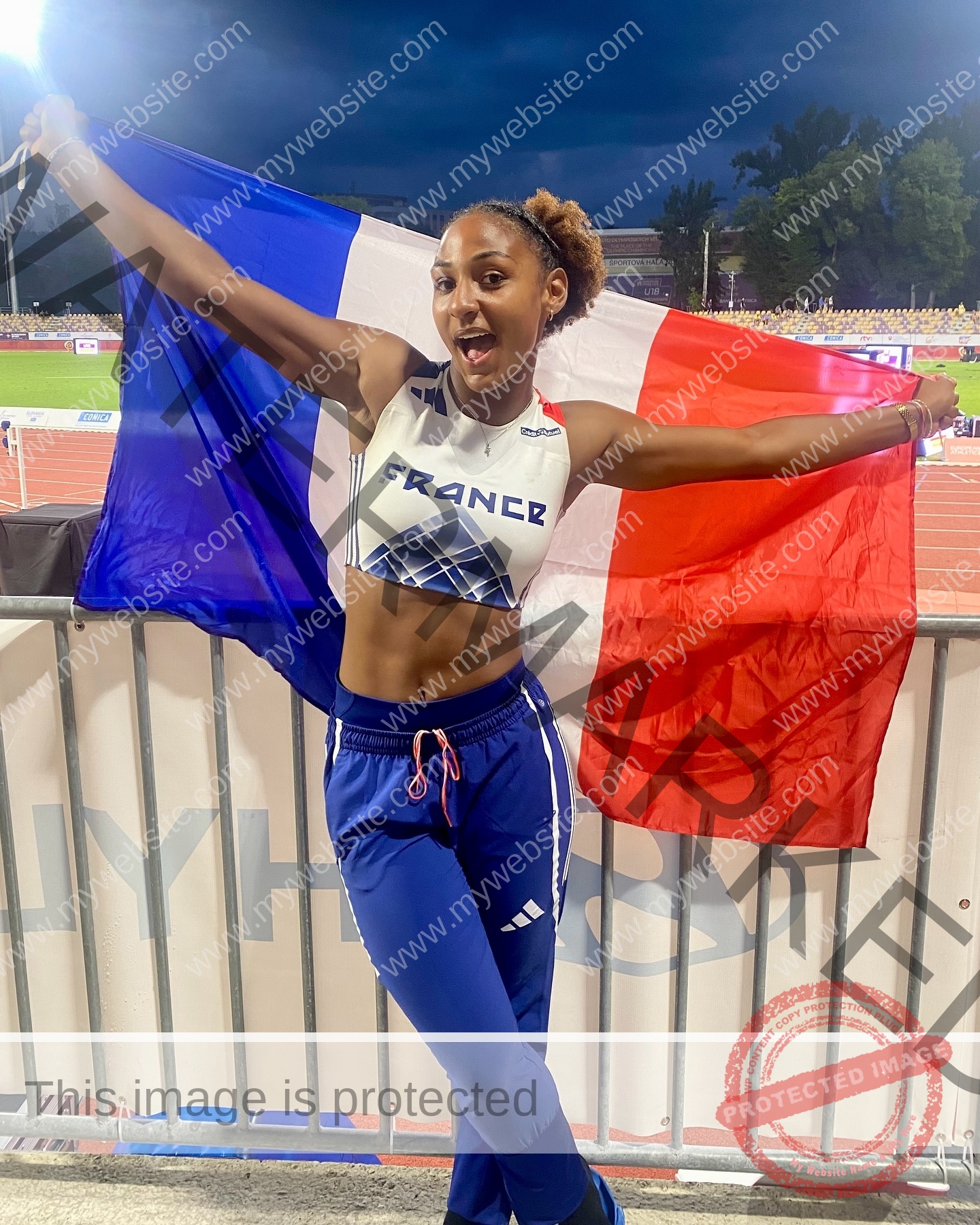 zola-ndouma-mona-france-zolaathle-09326 Zola Ndouma Mona, track and field star from France, wears a France crop top and blue pants, smiling while holding the French flag at twilight.