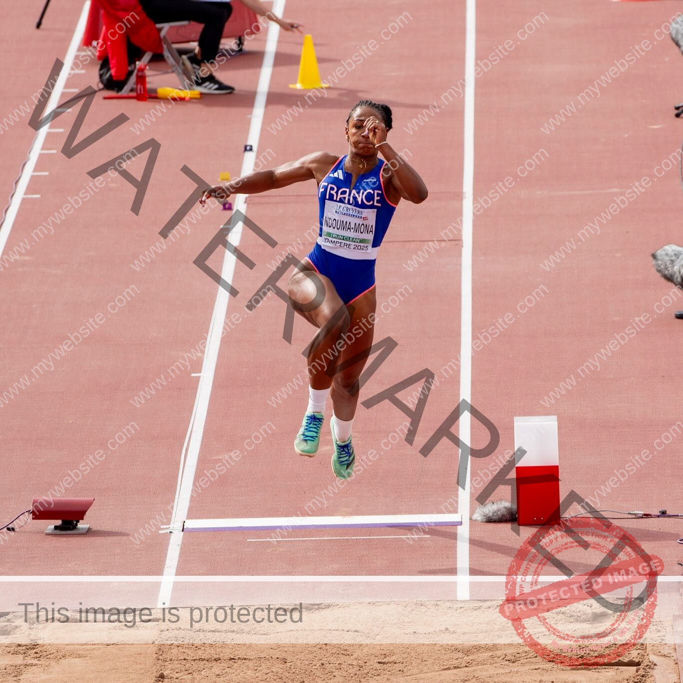 zola-ndouma-mona-france-zolaathle-09363 Zola Ndouma Mona, track and field star from France, mid-air in long jump at a meet, wearing blue and heading toward the sand pit.