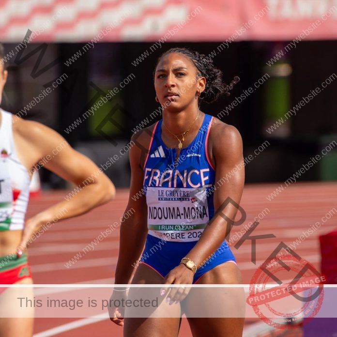 Zola Ndouma Mona, track and field star from France, stands on the track in blue uniform; another runner is partly seen on the left.