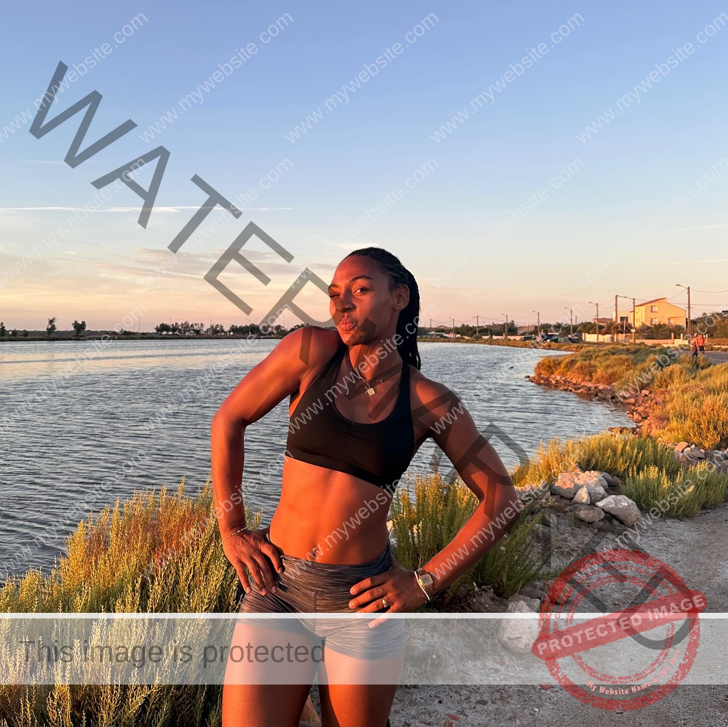 zola-ndouma-mona-france-zolaathle-09375 Zola Ndouma Mona, track and field star from France, poses confidently by water at sunset, smiling with hands on hips. Greenery behind.
