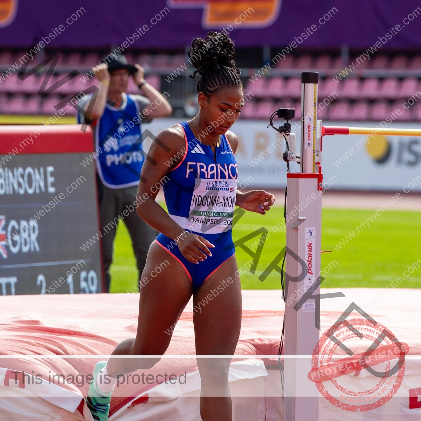 zola-ndouma-mona-france-zolaathle-09384 Zola Ndouma Mona, track and field star from France stands in a blue uniform by the high jump bar, ready to jump, crowd behind.